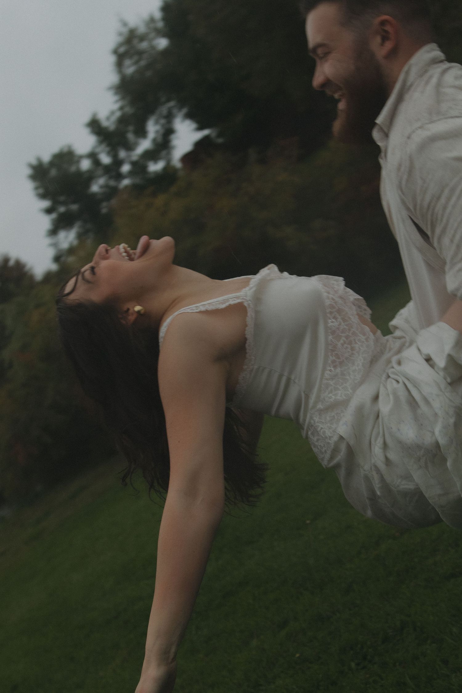 A romantic couple embraces outdoors in a grassy field at dusk wearing light-colored clothing.