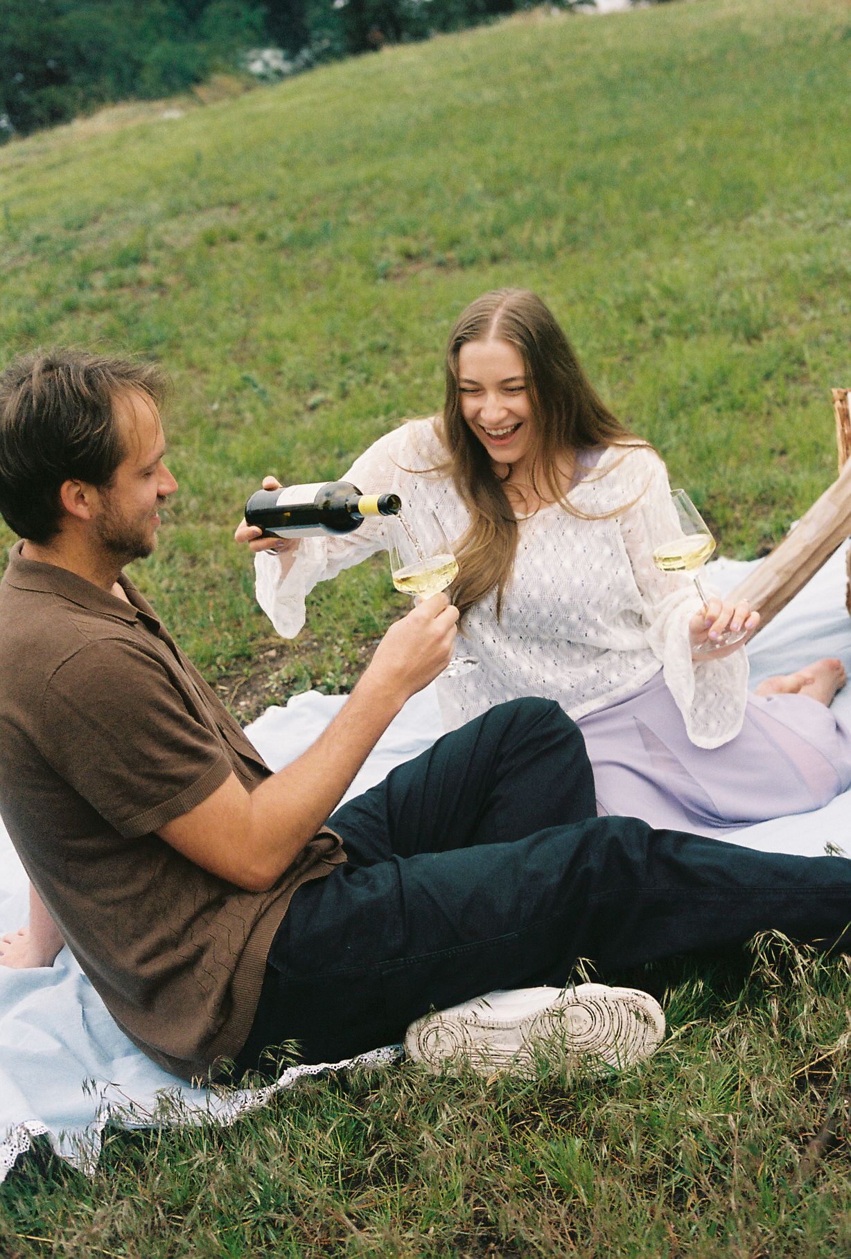 Couple laughing at a picnic as man pours white wine into a glass on a green grassy hillside.