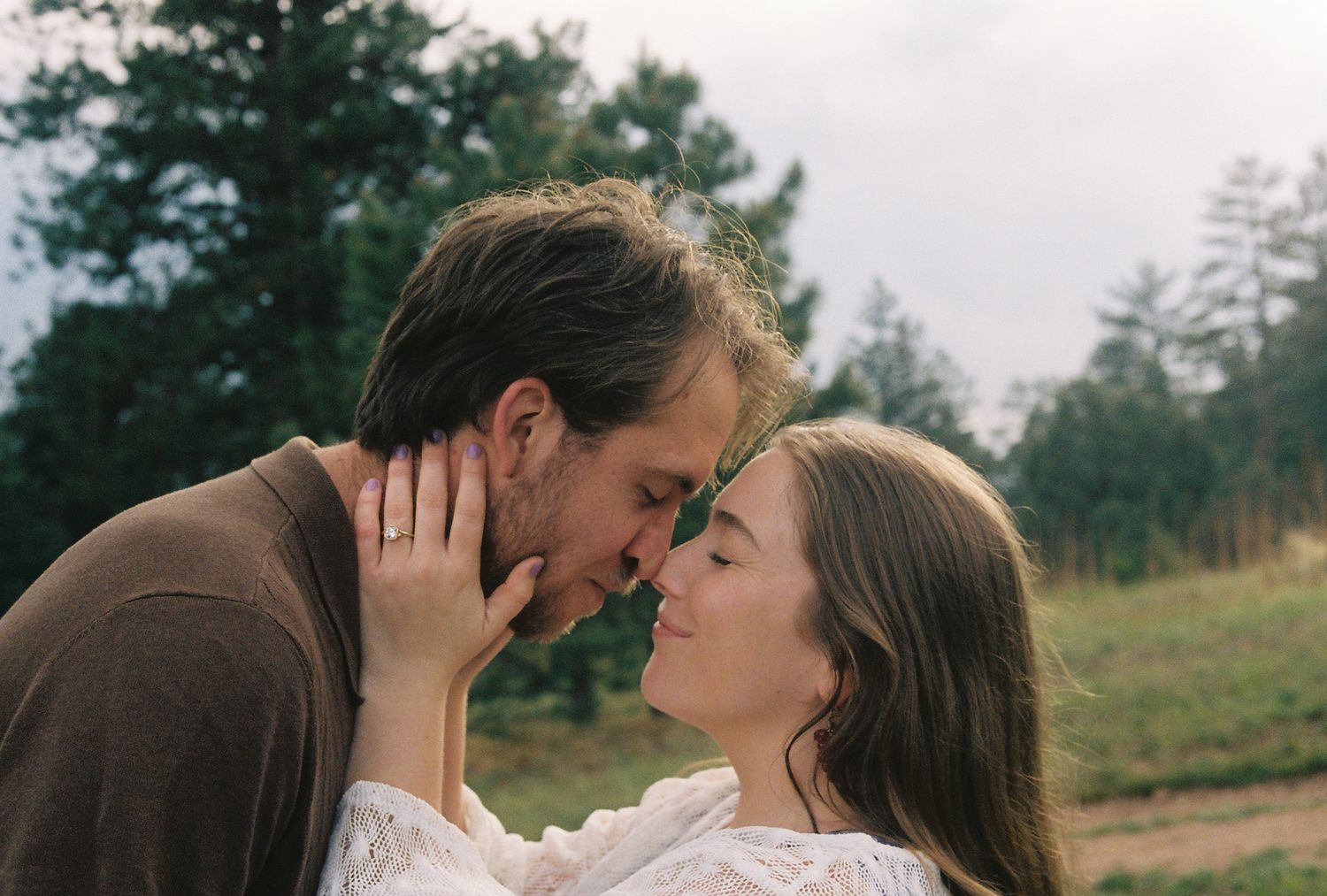 Couple touching foreheads in a tender romantic moment outdoors with green trees in background.