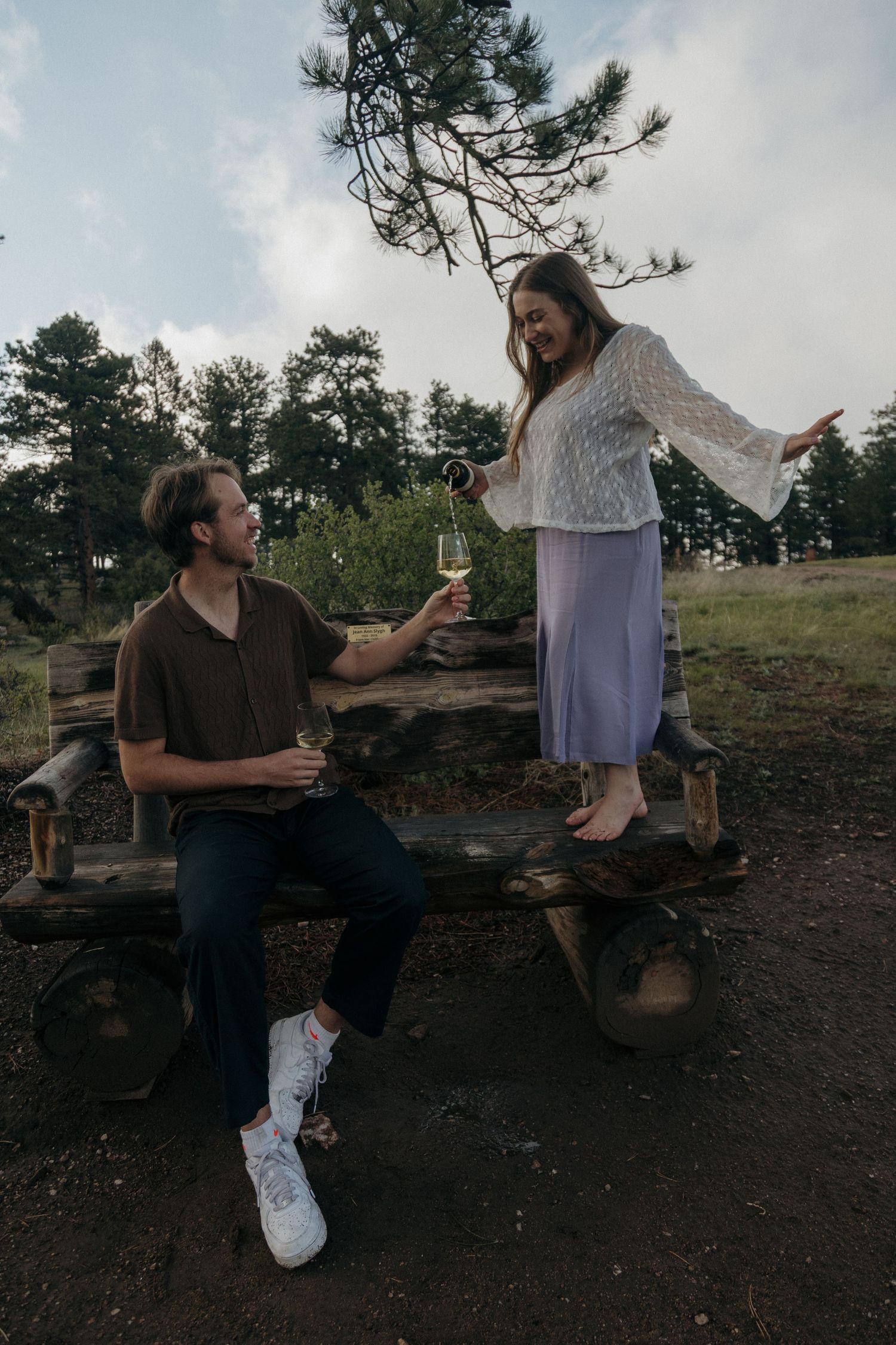Couple sharing wine glasses outdoors on a rustic wooden bench surrounded by pine trees at dusk.