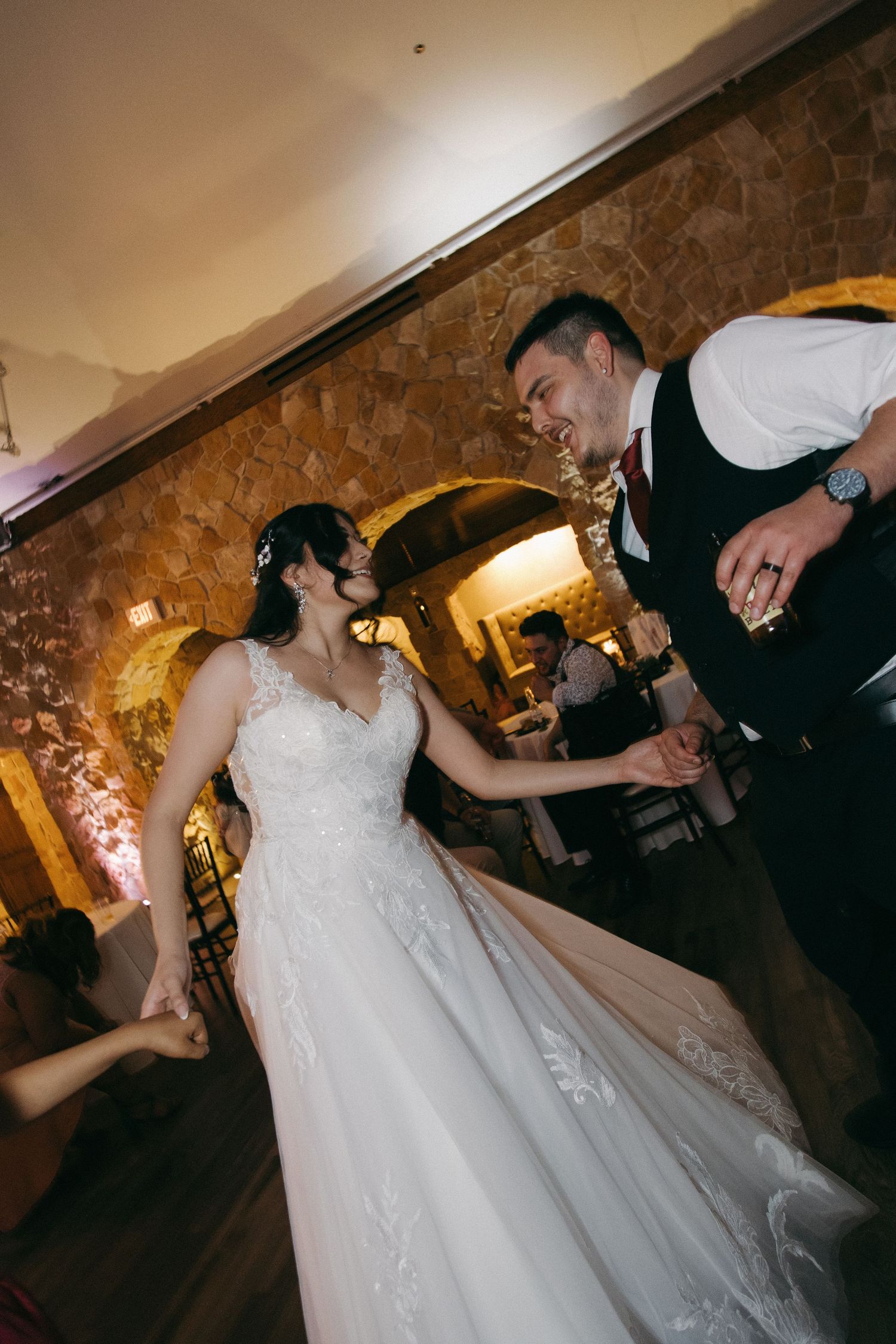 Bride and groom share a joyful first dance in a warmly lit stone-walled wedding reception venue.