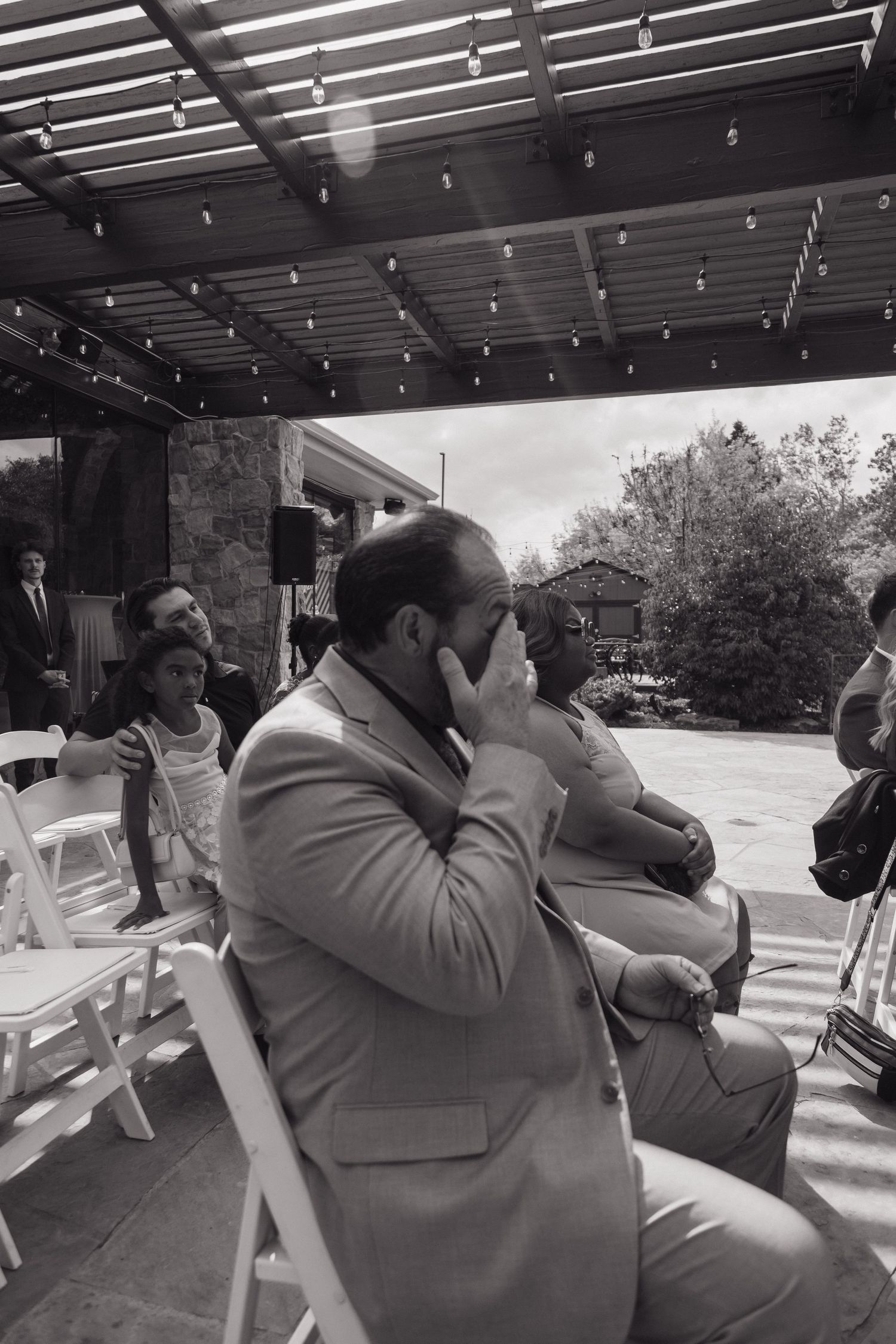Emotional wedding guest wiping tears during outdoor ceremony under pergola in black and white photo.