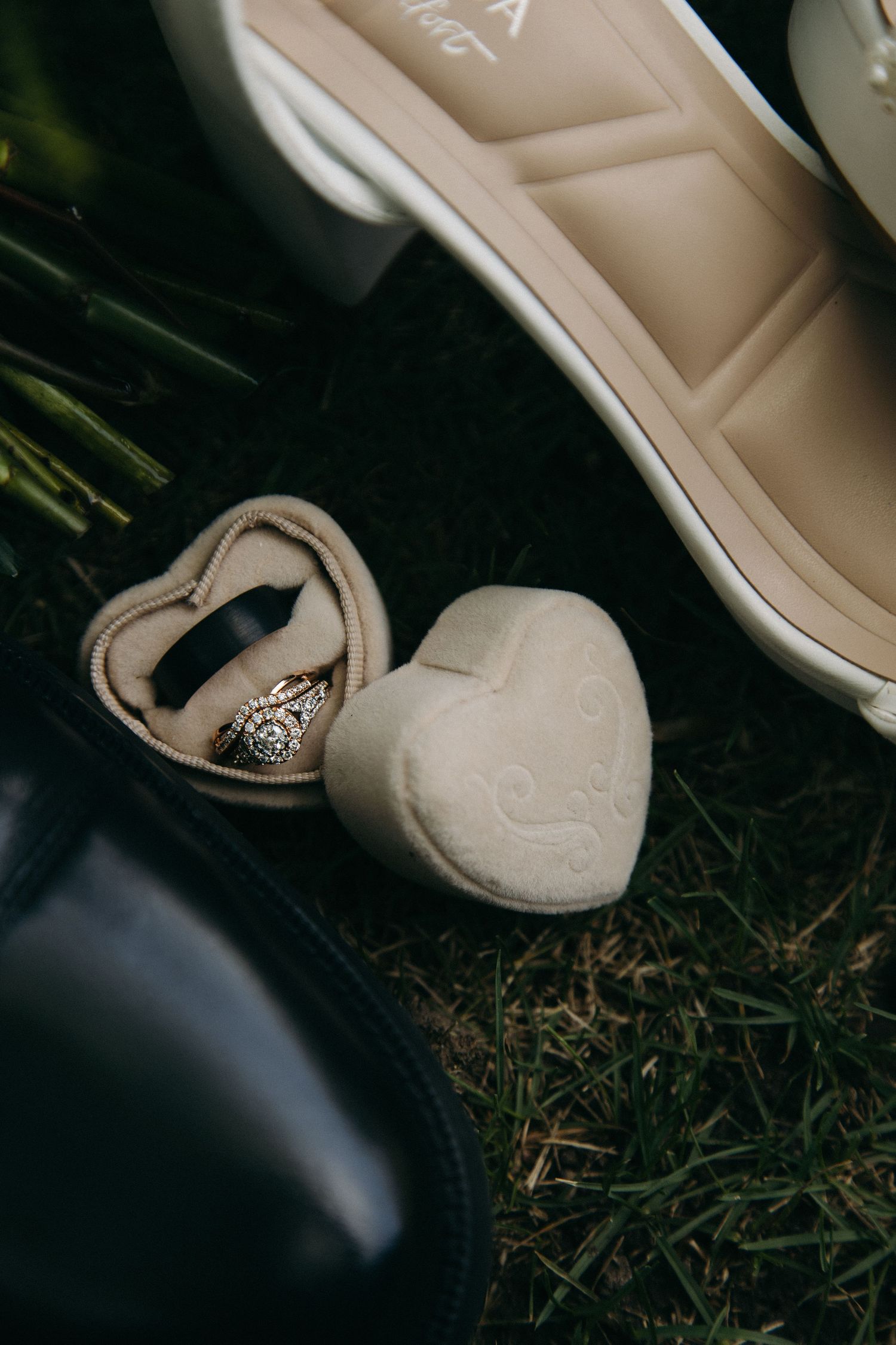 Heart-shaped velvet ring box with wedding rings on grass between bridal heels and groom dress shoes.