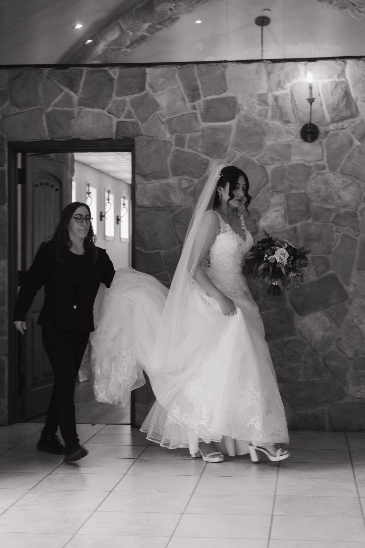 Bride in lace gown and veil enters stone chapel assisted by wedding coordinator in black.