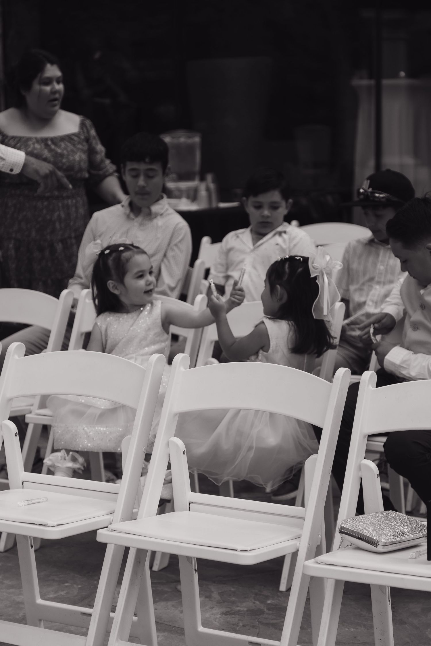 Black and white photo of flower girls and ring bearers seated on white chairs at a wedding ceremony.