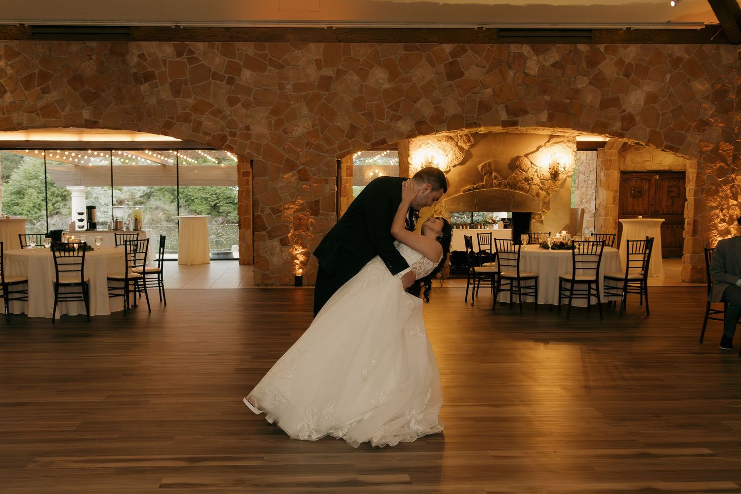 Groom dips bride in white gown during romantic first dance in stone-walled venue.