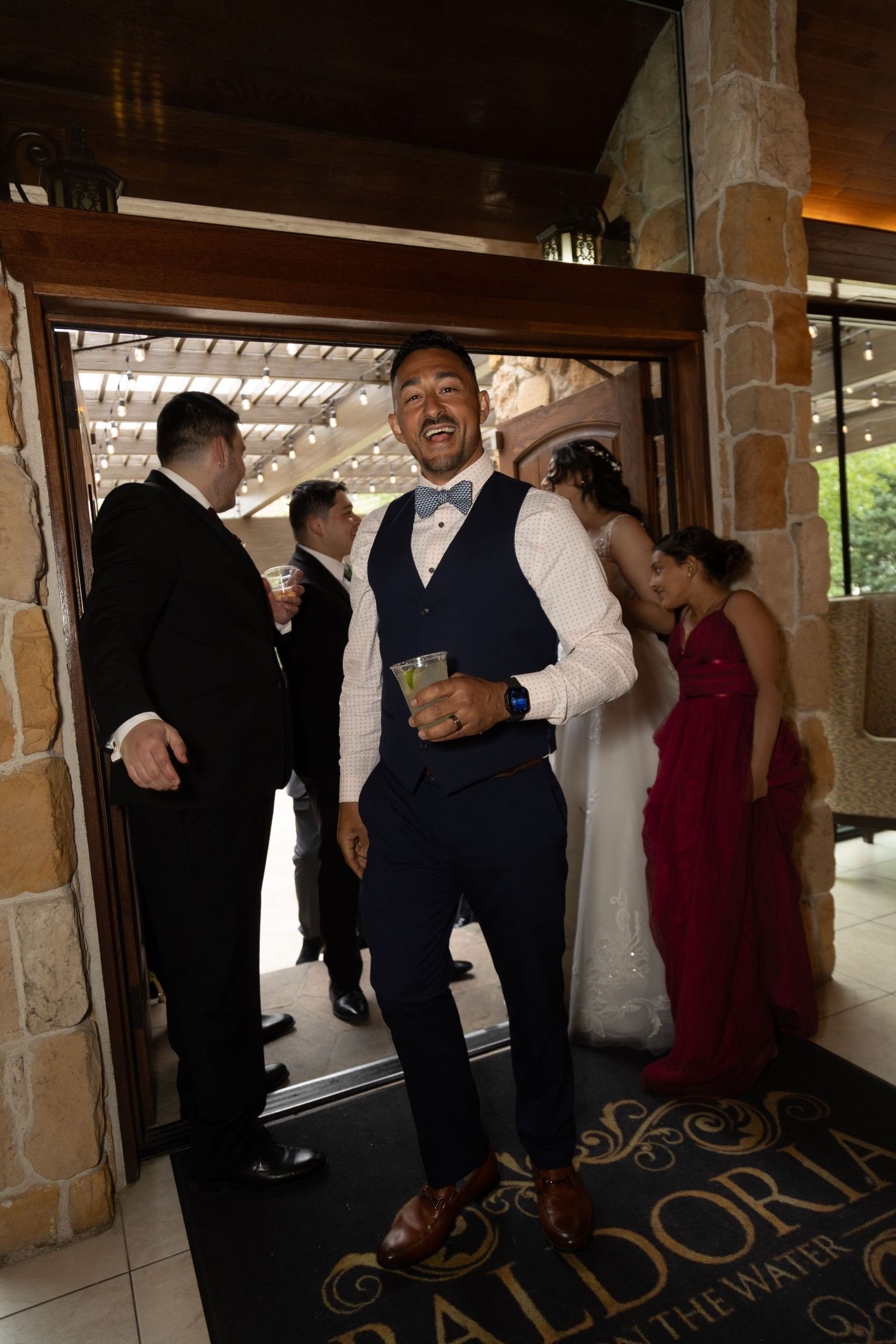A smiling groomsman in a navy vest holds a drink at the wedding venue entrance.