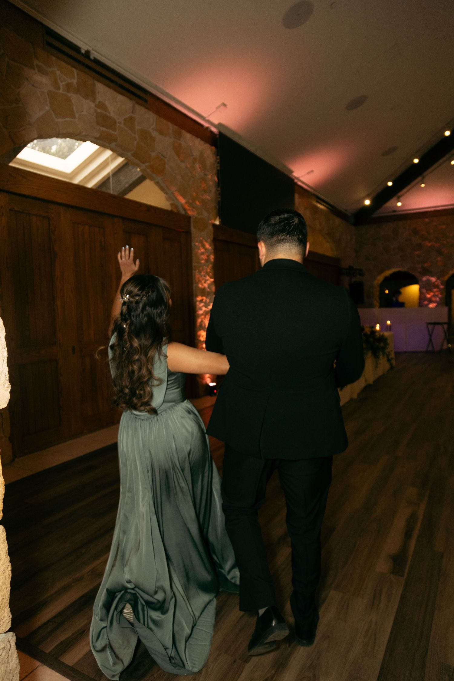 Couple in formal attire walking through a rustic stone venue, woman in sage green gown and man in black suit.