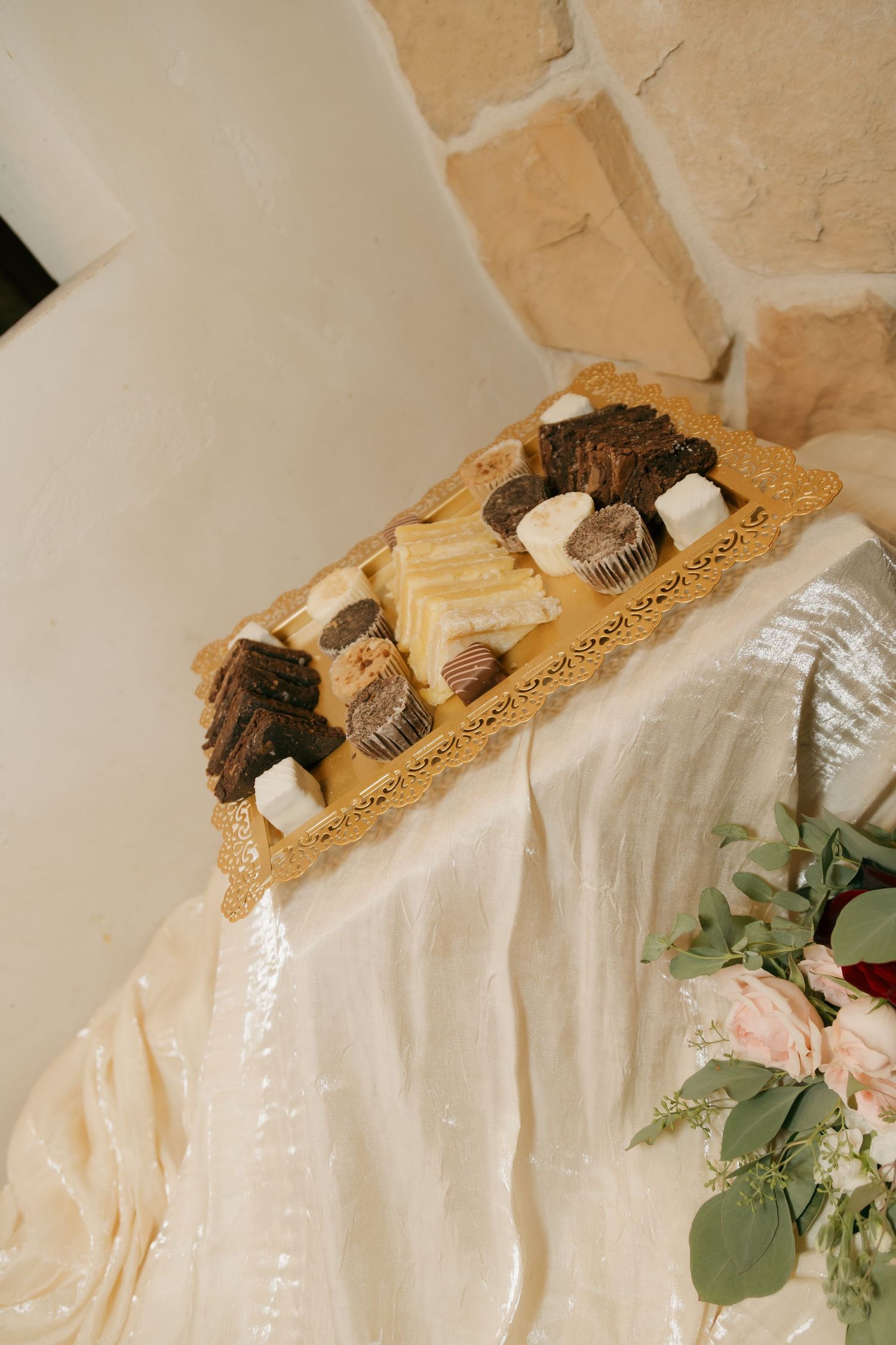 Assorted wedding desserts including brownies and chocolates on gold tray with floral arrangement nearby.