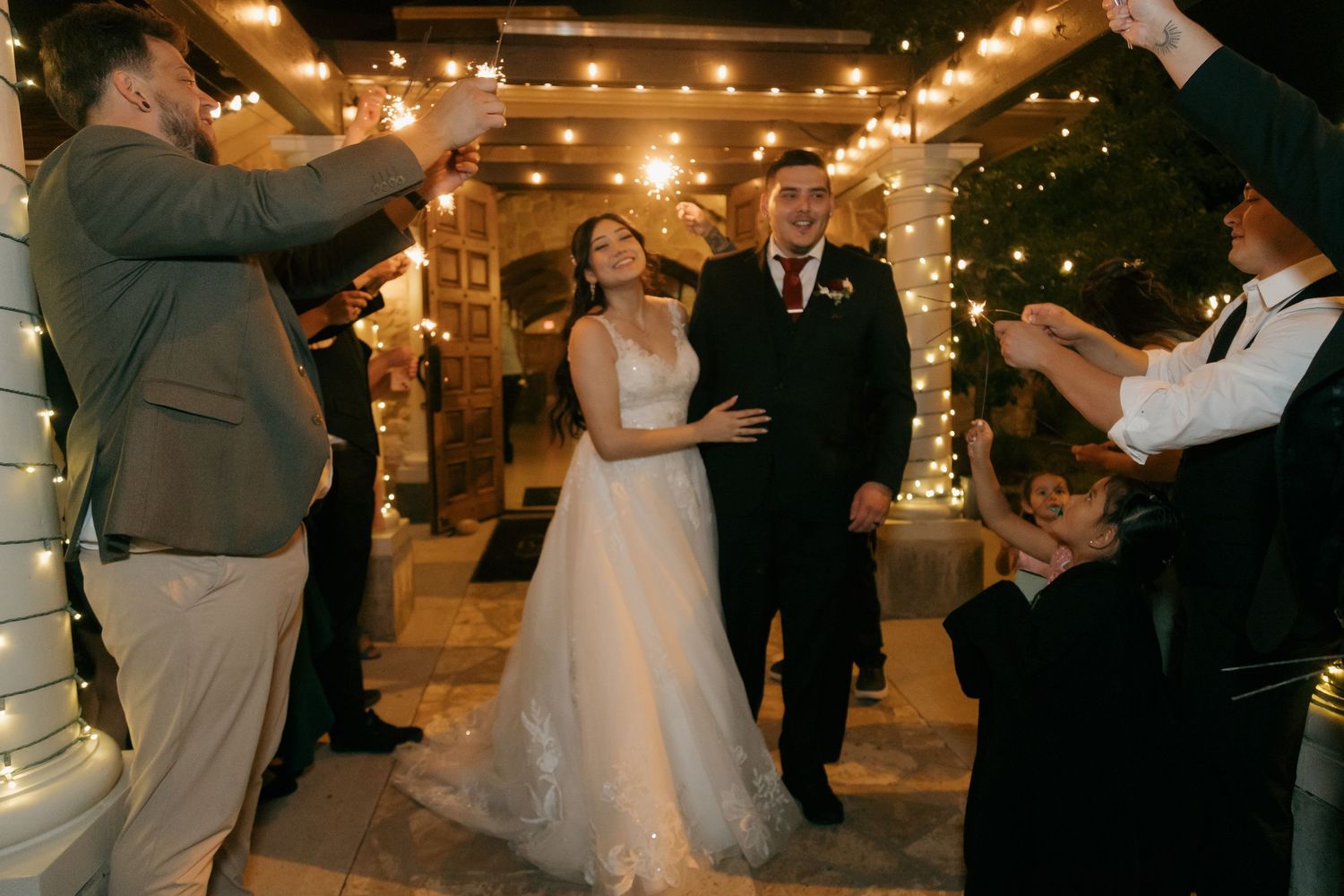 Bride and groom exit wedding reception through sparkler send-off tunnel surrounded by cheering guests at night.