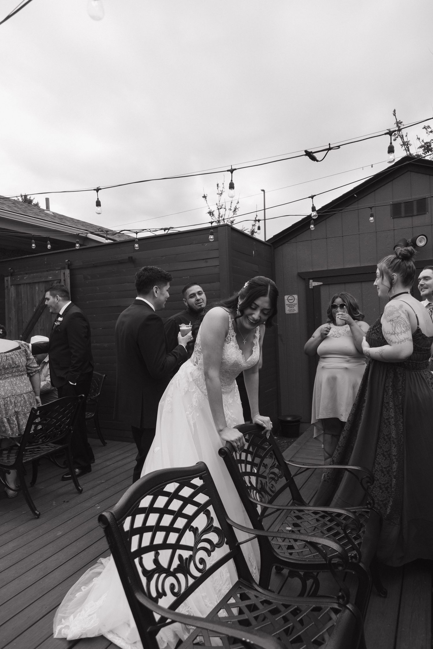 Black and white photo of bride adjusting chair on outdoor deck surrounded by wedding guests.