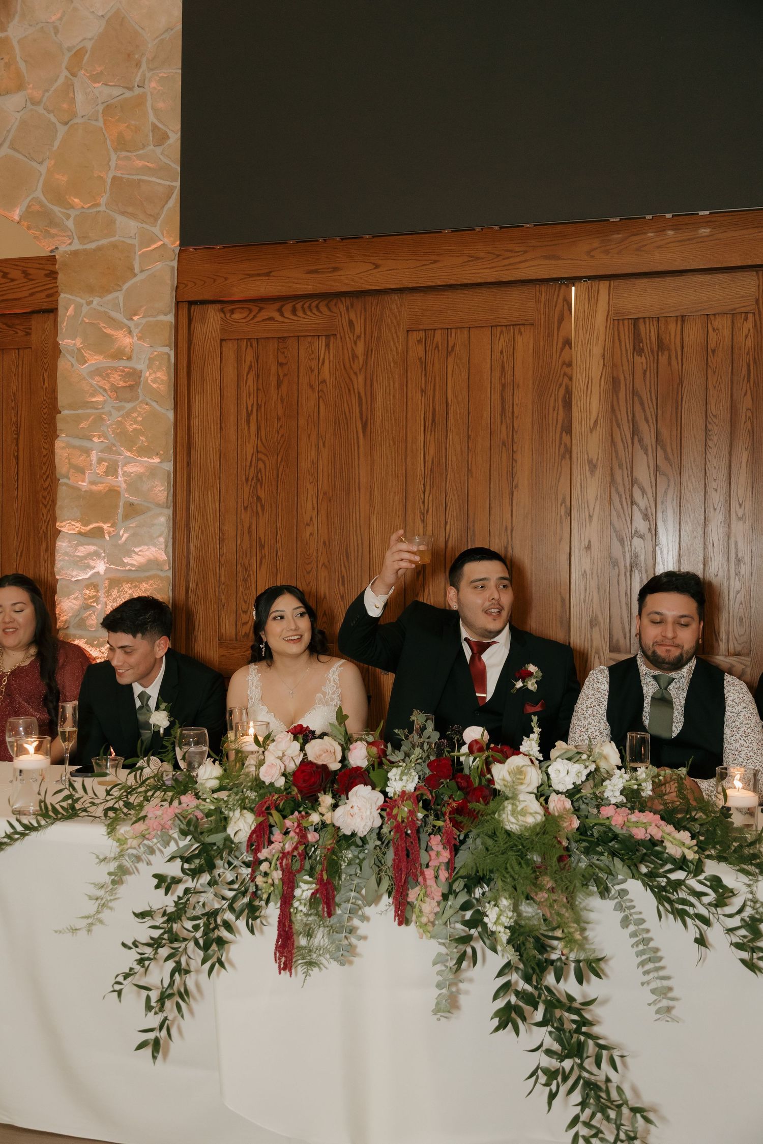 Bride and groom raise a toast at the head table adorned with lush floral arrangements at reception.