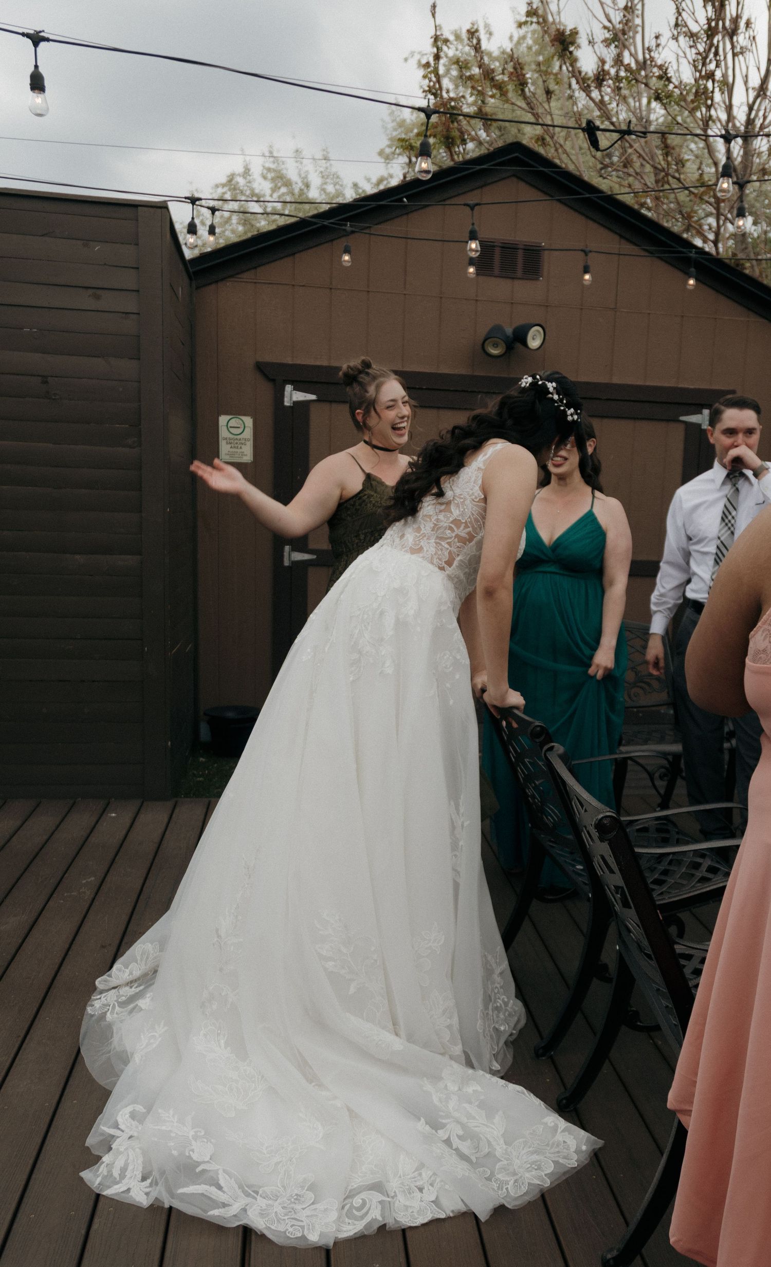 Bridesmaids in emerald green dresses assist bride with her lace wedding gown on an outdoor deck.