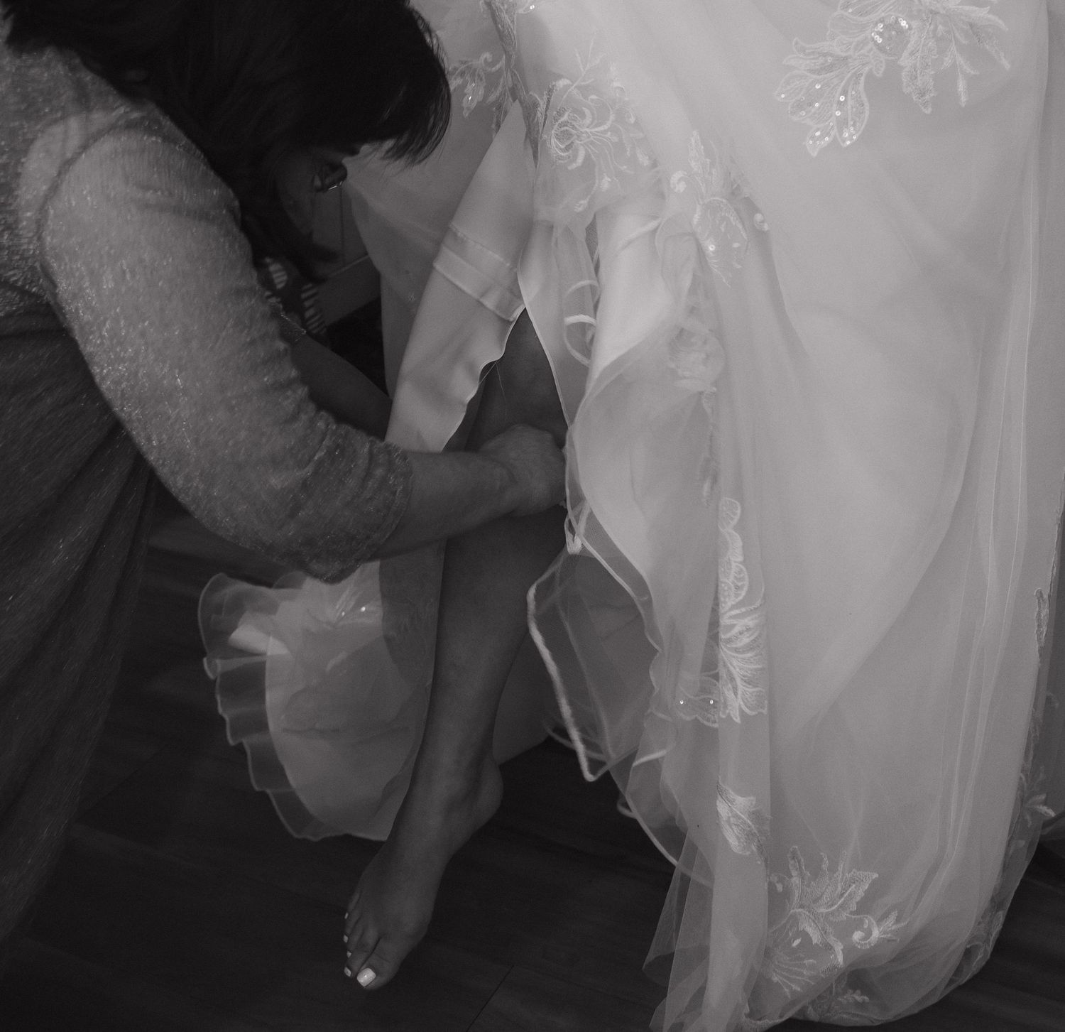Black and white photo of bridesmaid helping bride with shoes before wedding.