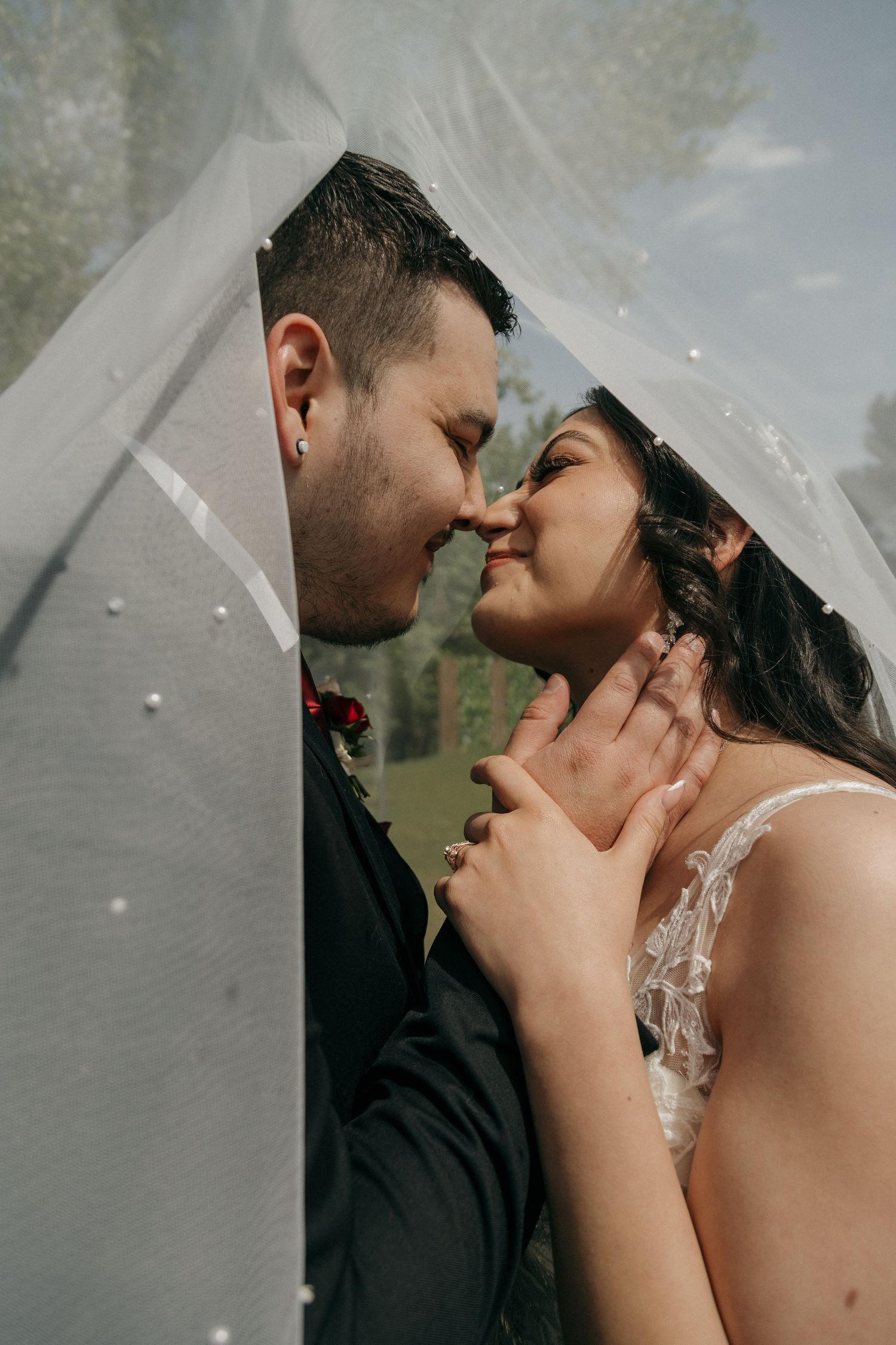Bride and groom share a romantic almost-kiss under a pearl-adorned wedding veil outdoors.