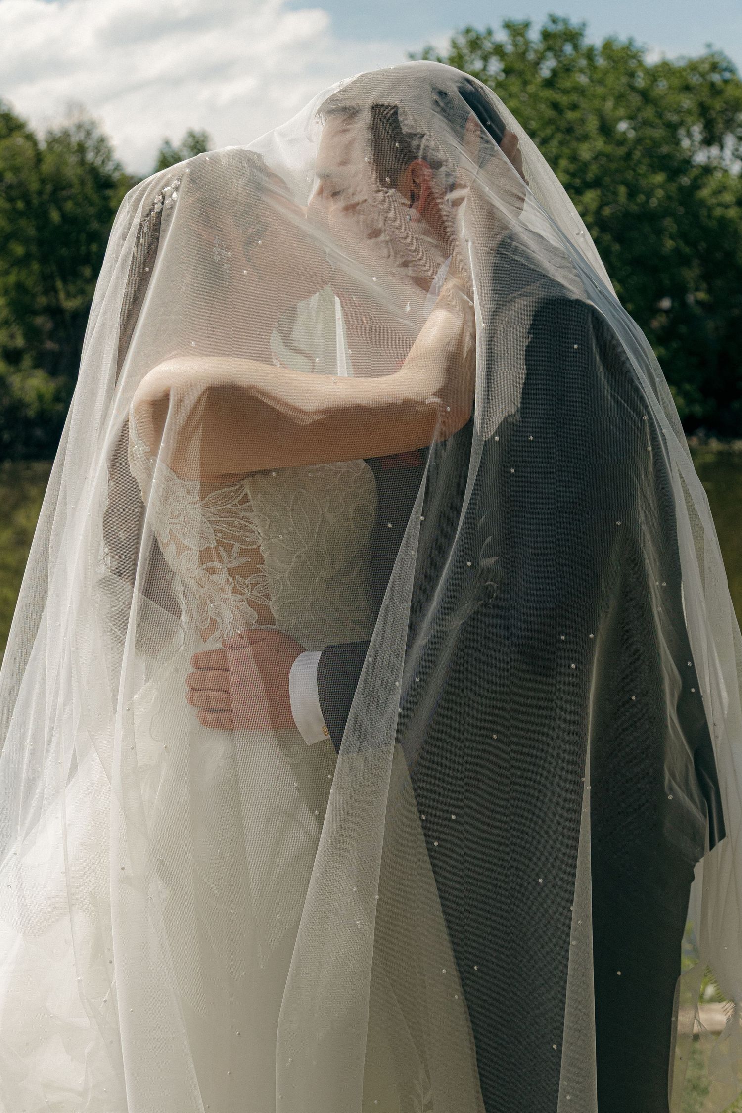 Bride and groom share an intimate kiss beneath a flowing pearl-adorned wedding veil outdoors.