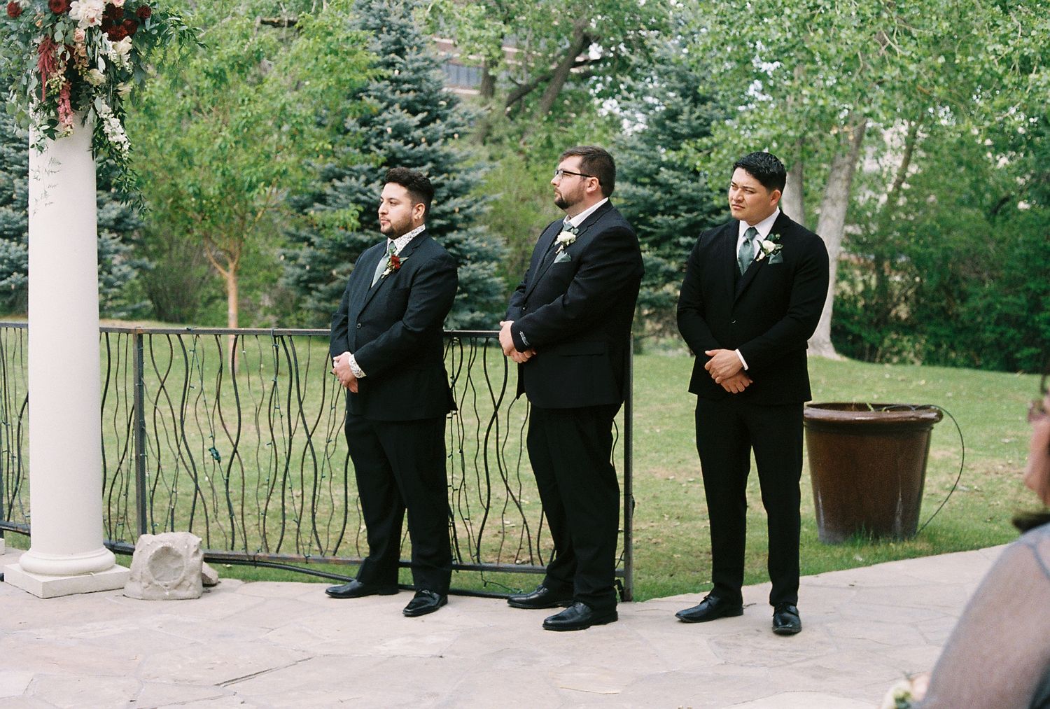 Three groomsmen in black suits stand at attention during outdoor wedding ceremony.