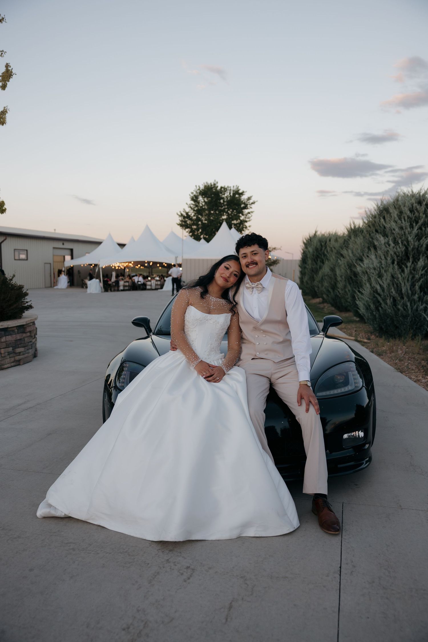 A bride and groom pose together leaning on a black sports car at their outdoor evening wedding reception.