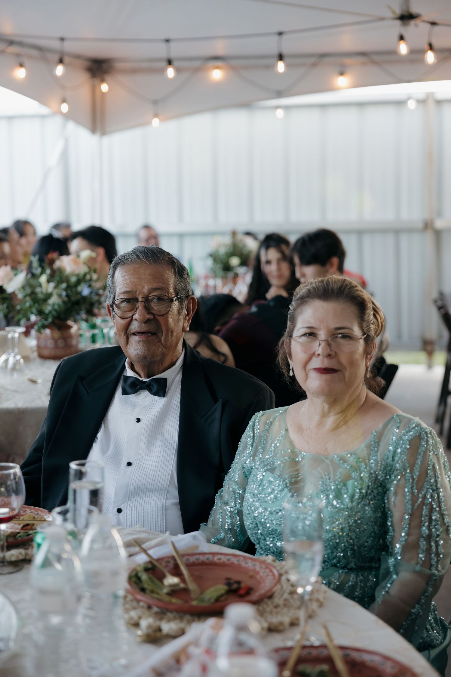 Elderly couple in formal attire smiling at a wedding reception under string lights.
