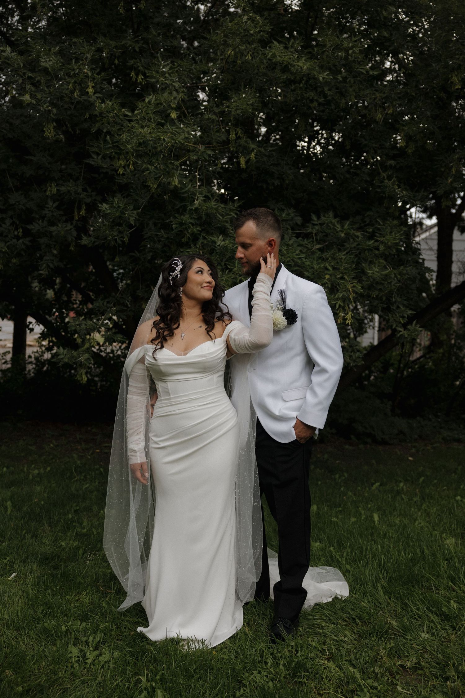 Bride in white gown with veil and groom in white jacket share a romantic moment outdoors on green grass.