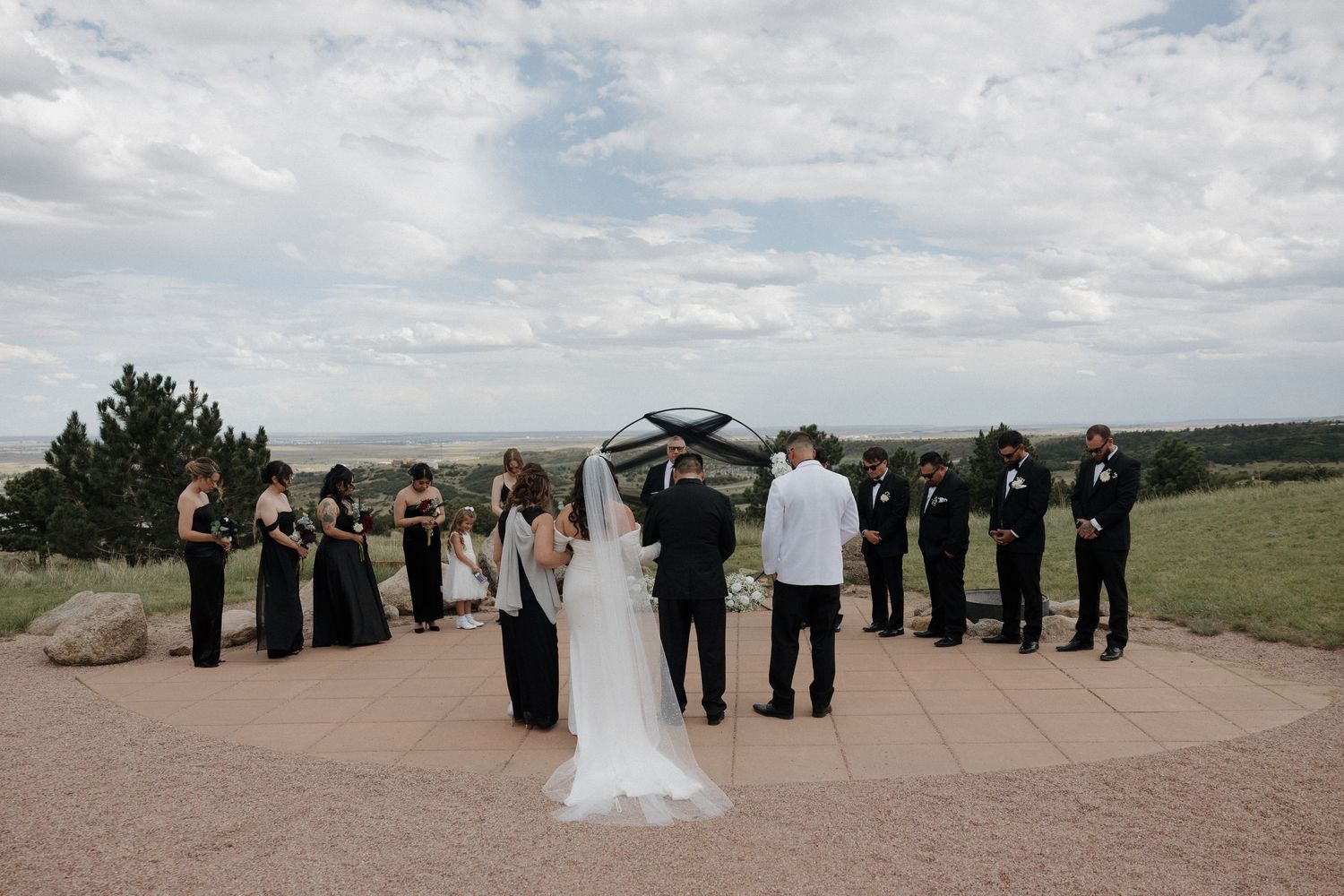 Outdoor wedding ceremony on a hilltop patio with bride, groom, officiant, bridesmaids in black, and groomsmen under cloudy skies.