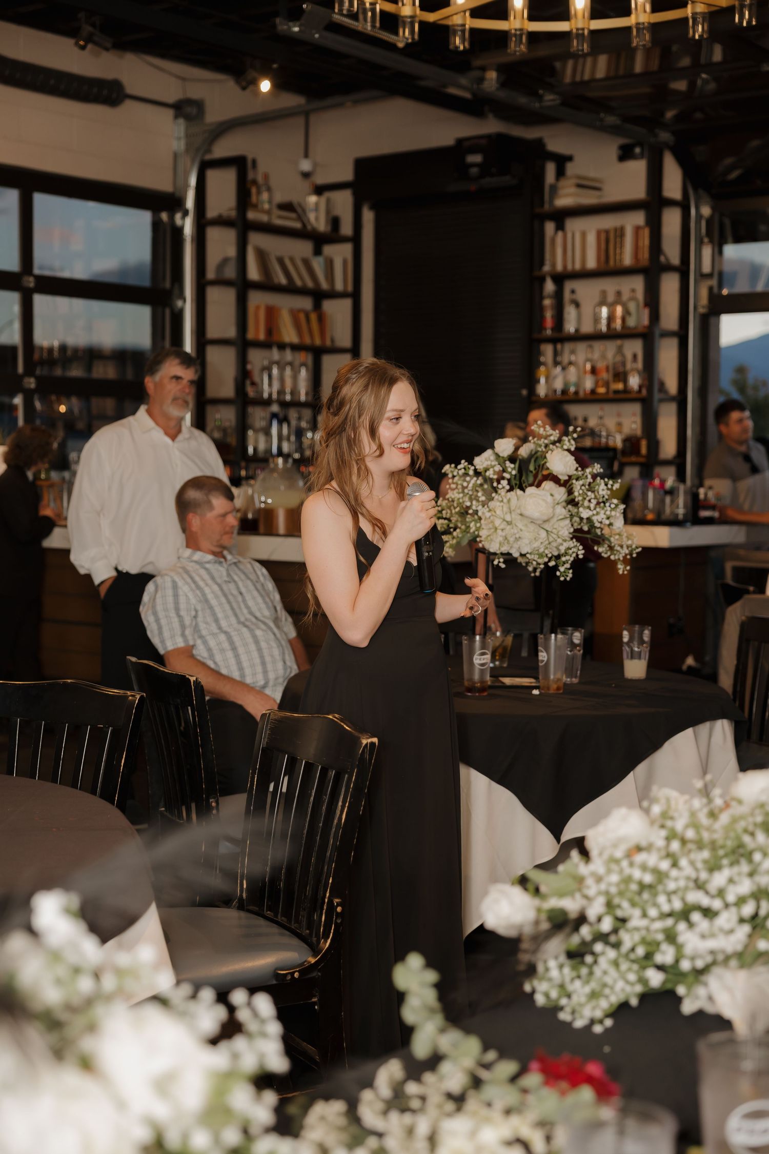 Bridesmaid in black gown gives speech at elegant indoor wedding reception with bar backdrop.