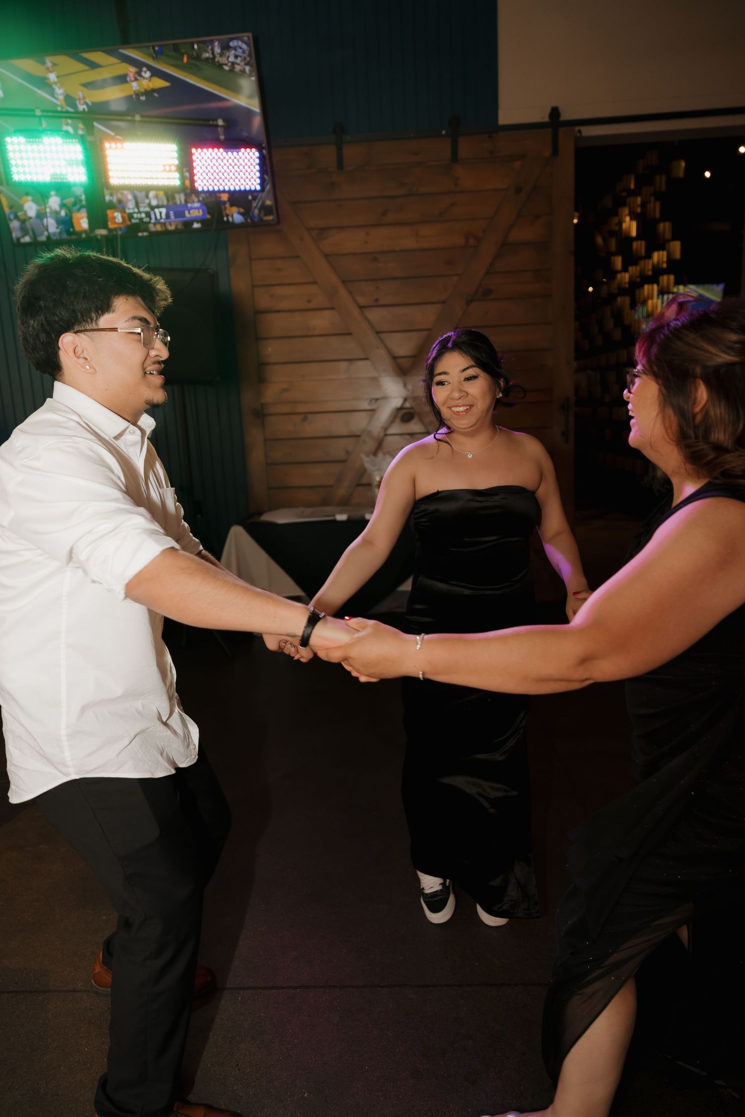 Guests dancing and holding hands at indoor wedding reception with rustic barn wood backdrop.