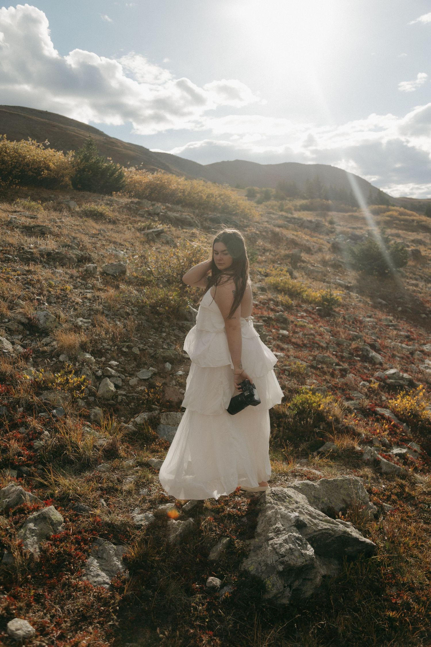 Bride in white tiered gown standing on rocky mountain terrain with sun flare and clouds in background.