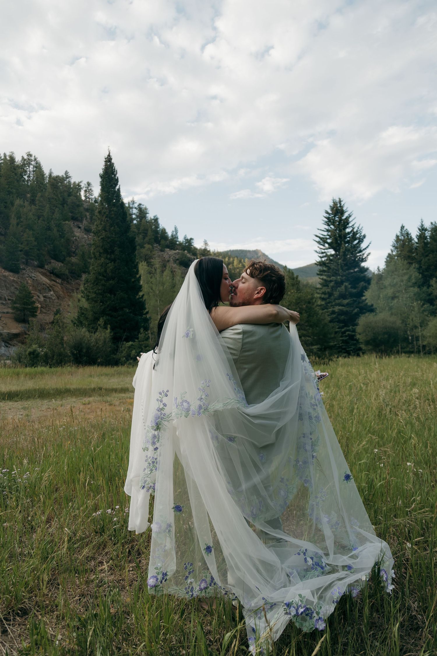 Groom lifts bride in floral embroidered veil as they share a kiss in a scenic mountain meadow landscape.