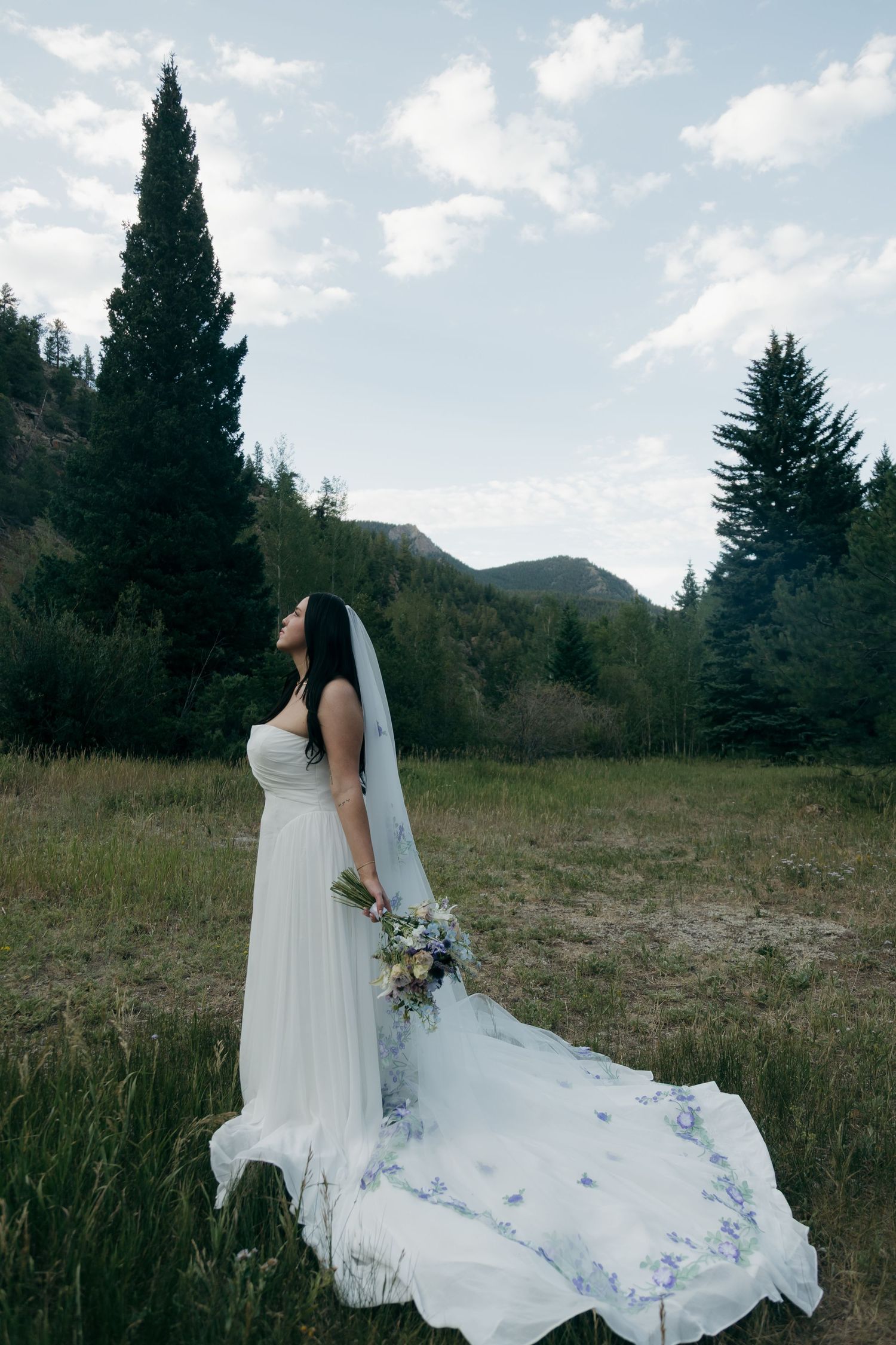 Bride in white gown with floral veil holds bouquet in mountain meadow surrounded by evergreen trees.
