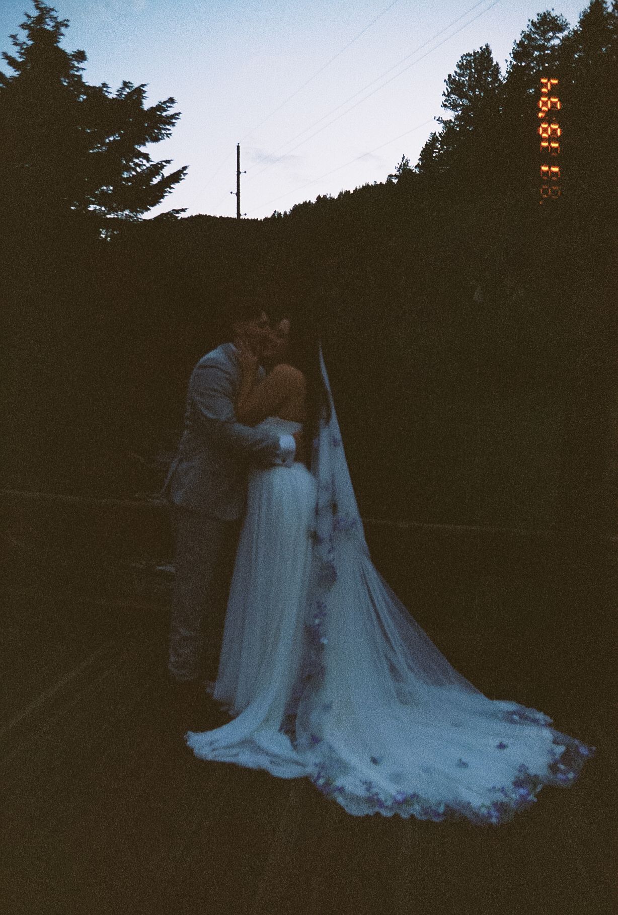 Silhouetted bride and groom share a romantic kiss at dusk, brides long embroidered veil trailing behind in a film photo.