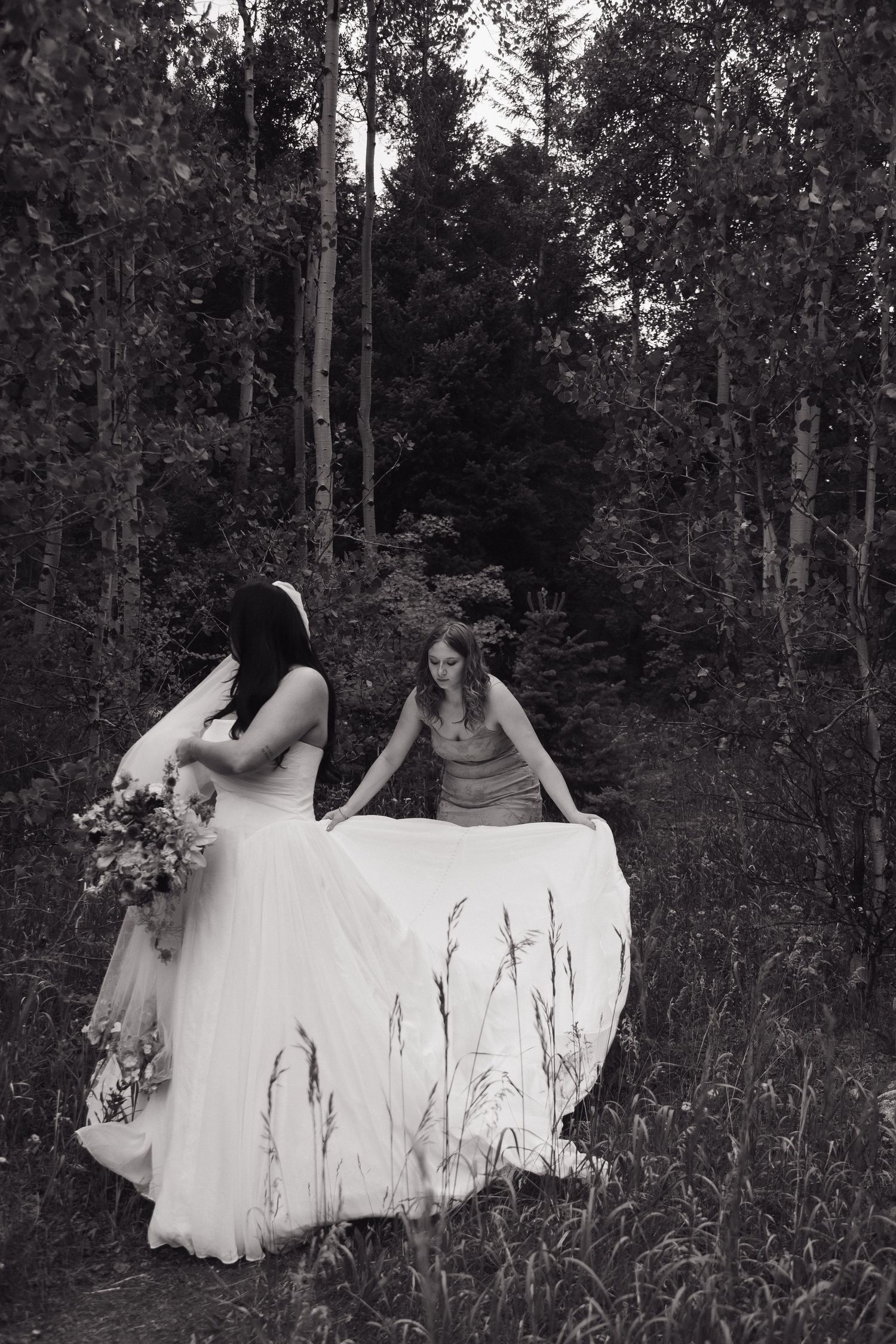 Black and white photo of bride holding bouquet as bridesmaid lifts wedding gown train in lush forest setting.