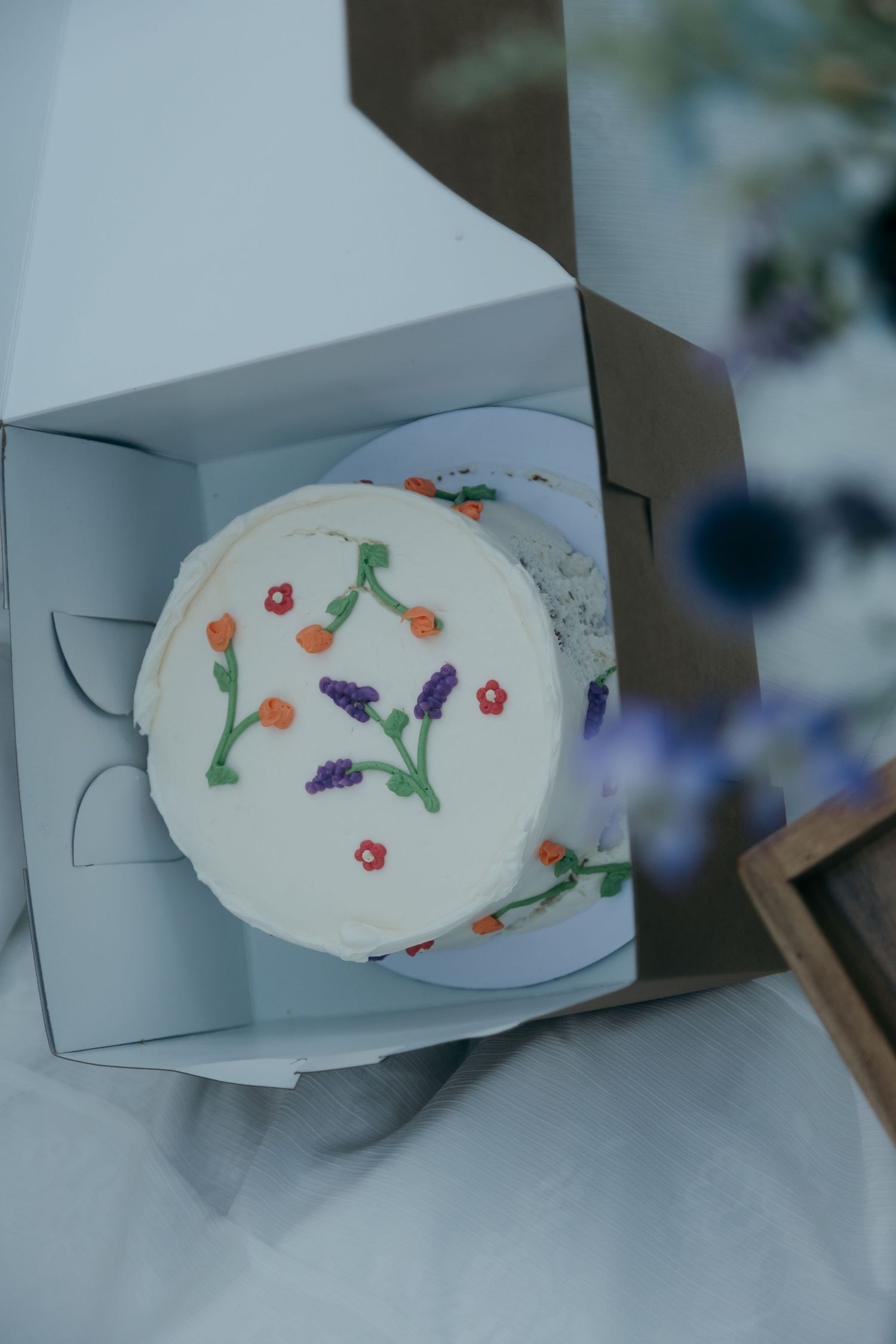 Elegant small white wedding cake decorated with colorful hand-piped floral details sits inside a white gift box.