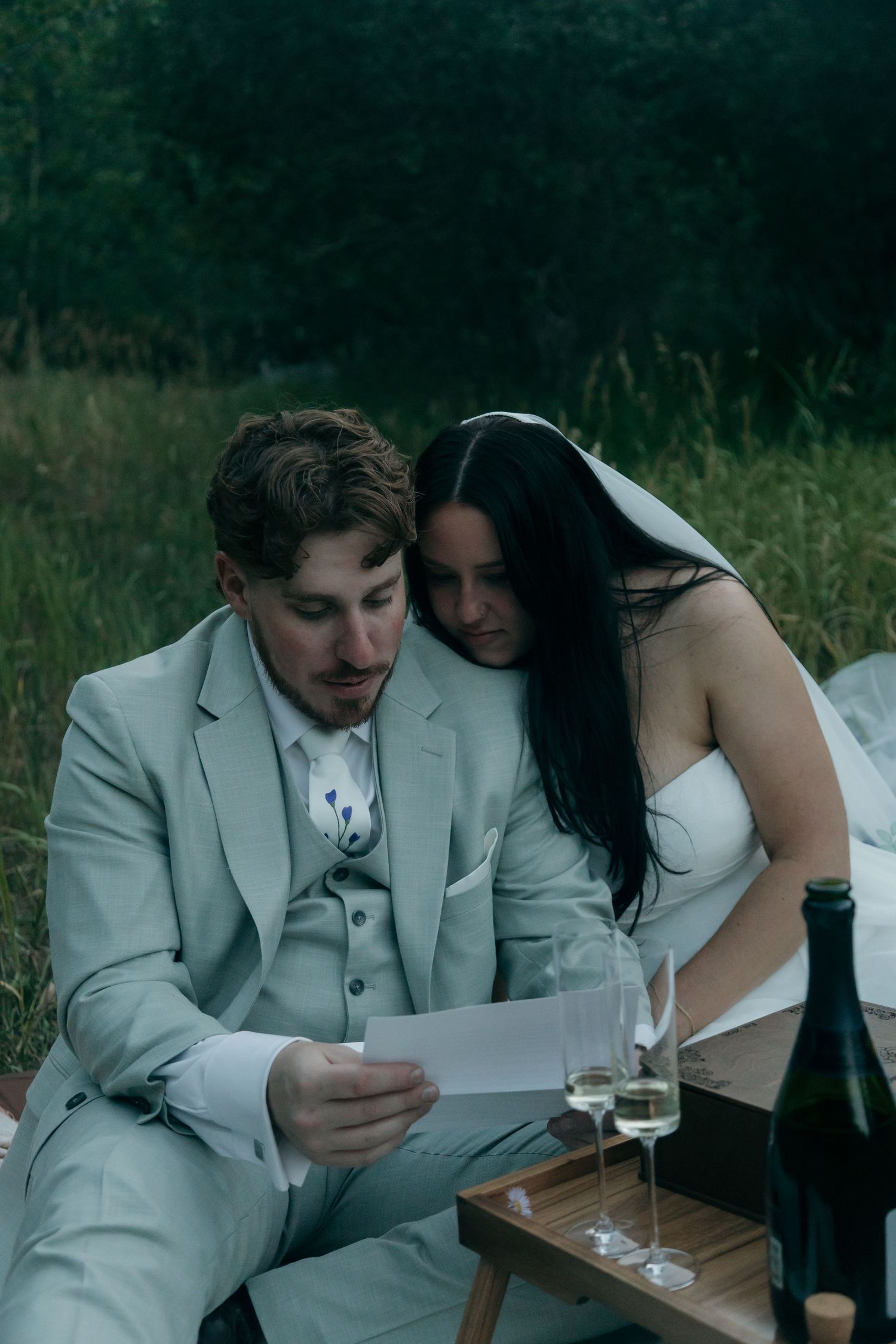 Bride leans on groom as he reads a heartfelt letter beside champagne glasses during their outdoor wedding picnic.