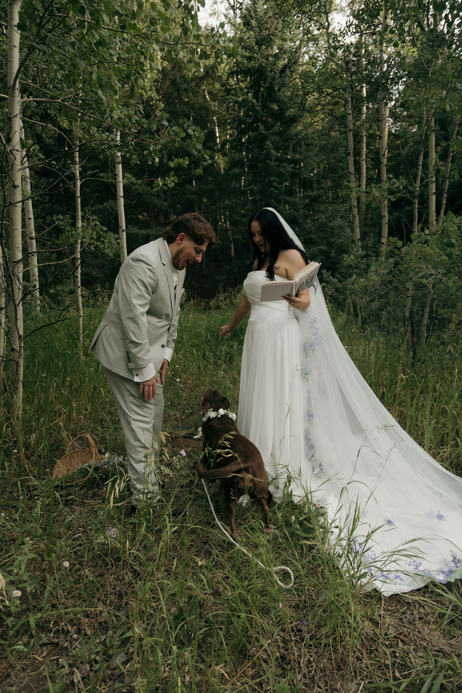 Bride reads vows in white gown with floral veil as groom and dog in flower crown listen in forest.