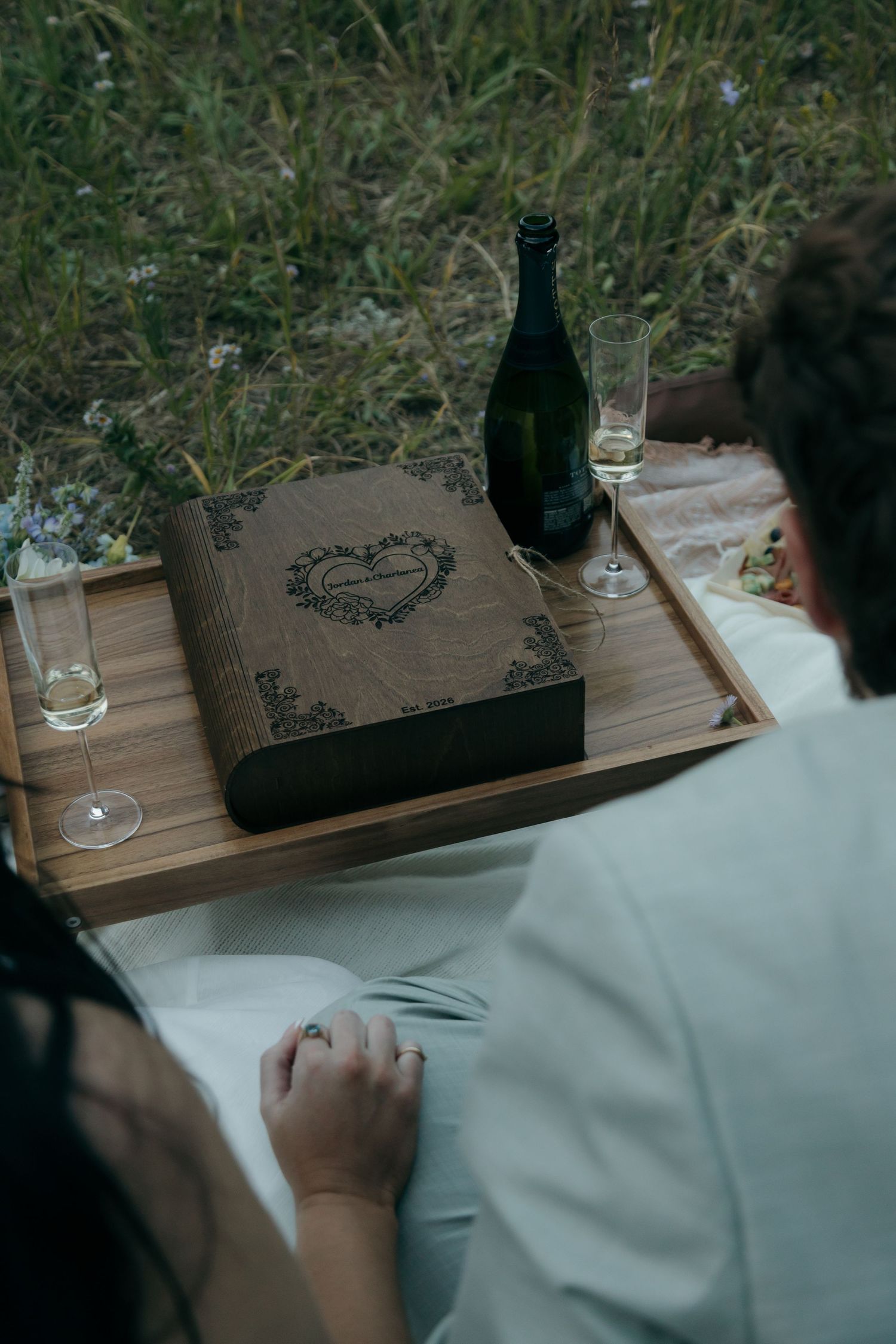 Elegant wooden engraved wedding box with champagne bottle and glasses on a wooden tray during an outdoor picnic celebration.
