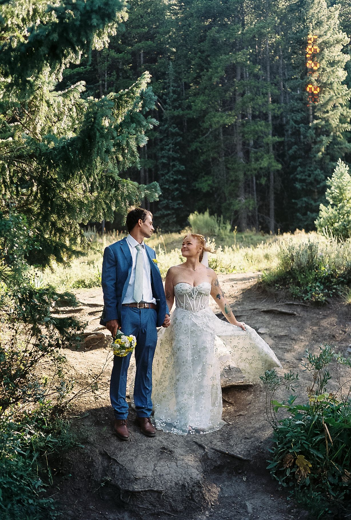 Bride in a strapless lace gown sits on a rock as groom stands beside her in a sunlit pine forest.
