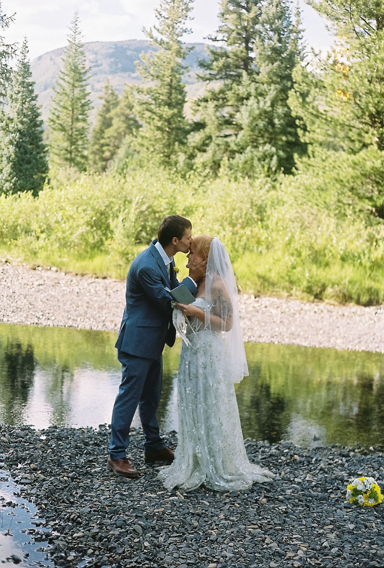 Bride and groom share first kiss by a mountain stream surrounded by lush green trees in an outdoor wedding ceremony.