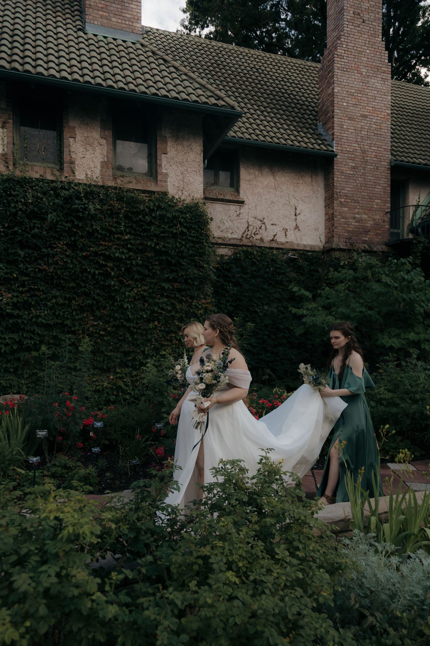 Bride in white gown with bridesmaids in green dresses holding bouquets in a lush garden beside a brick building.