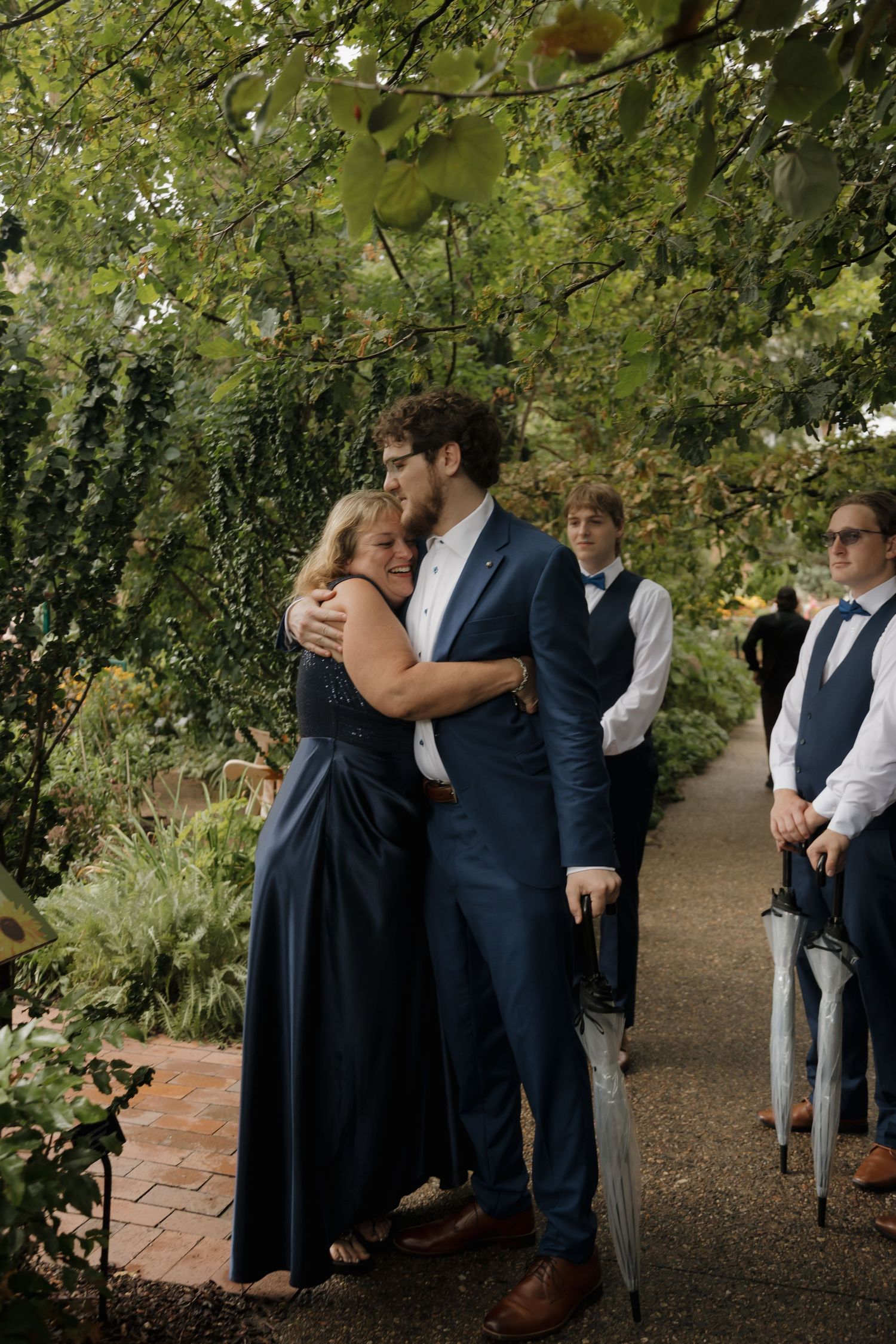 Groom in blue suit embracing his mother during outdoor wedding ceremony in lush garden setting.