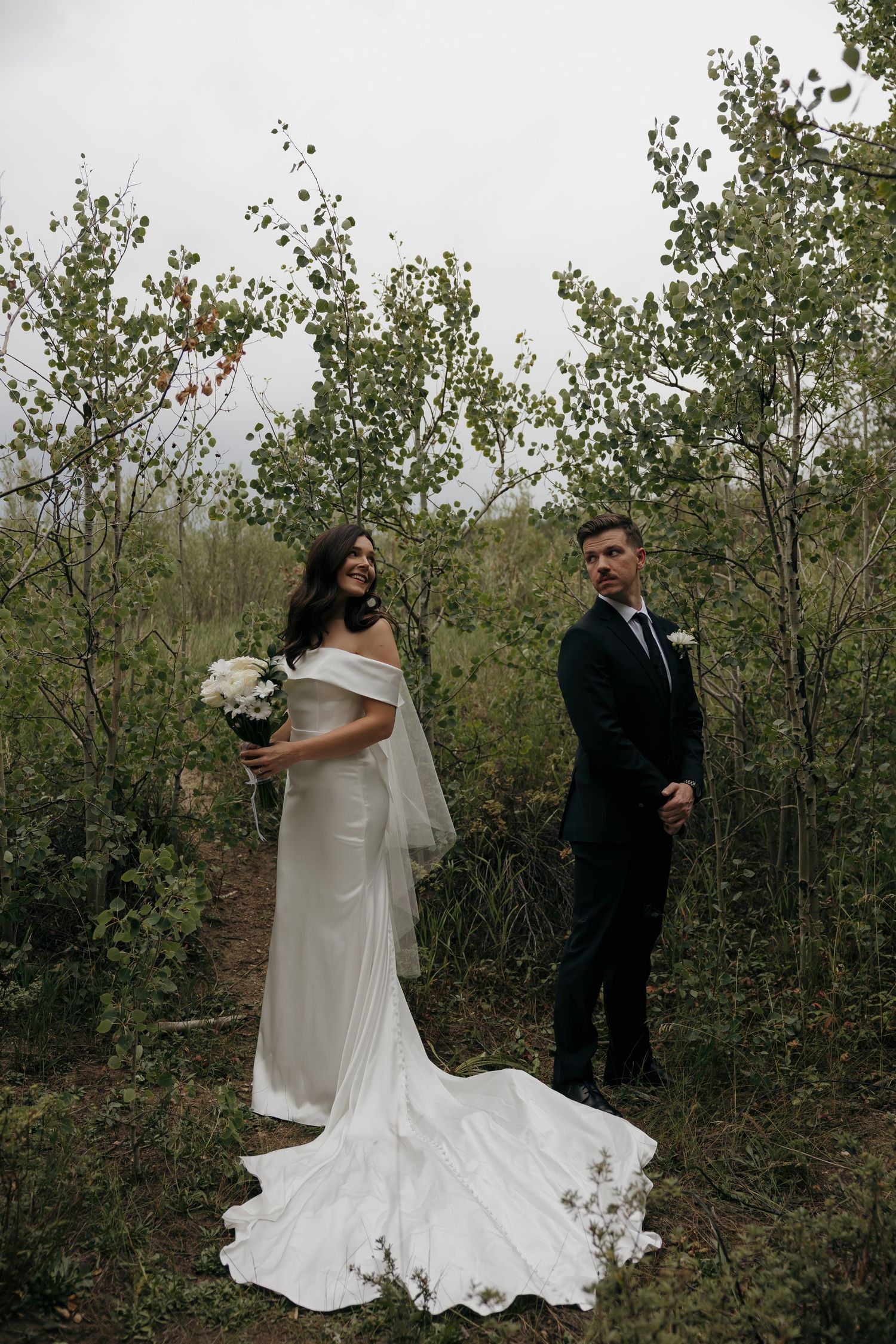 Bride and groom standing among aspen trees, bride in off-shoulder gown holding white bouquet.