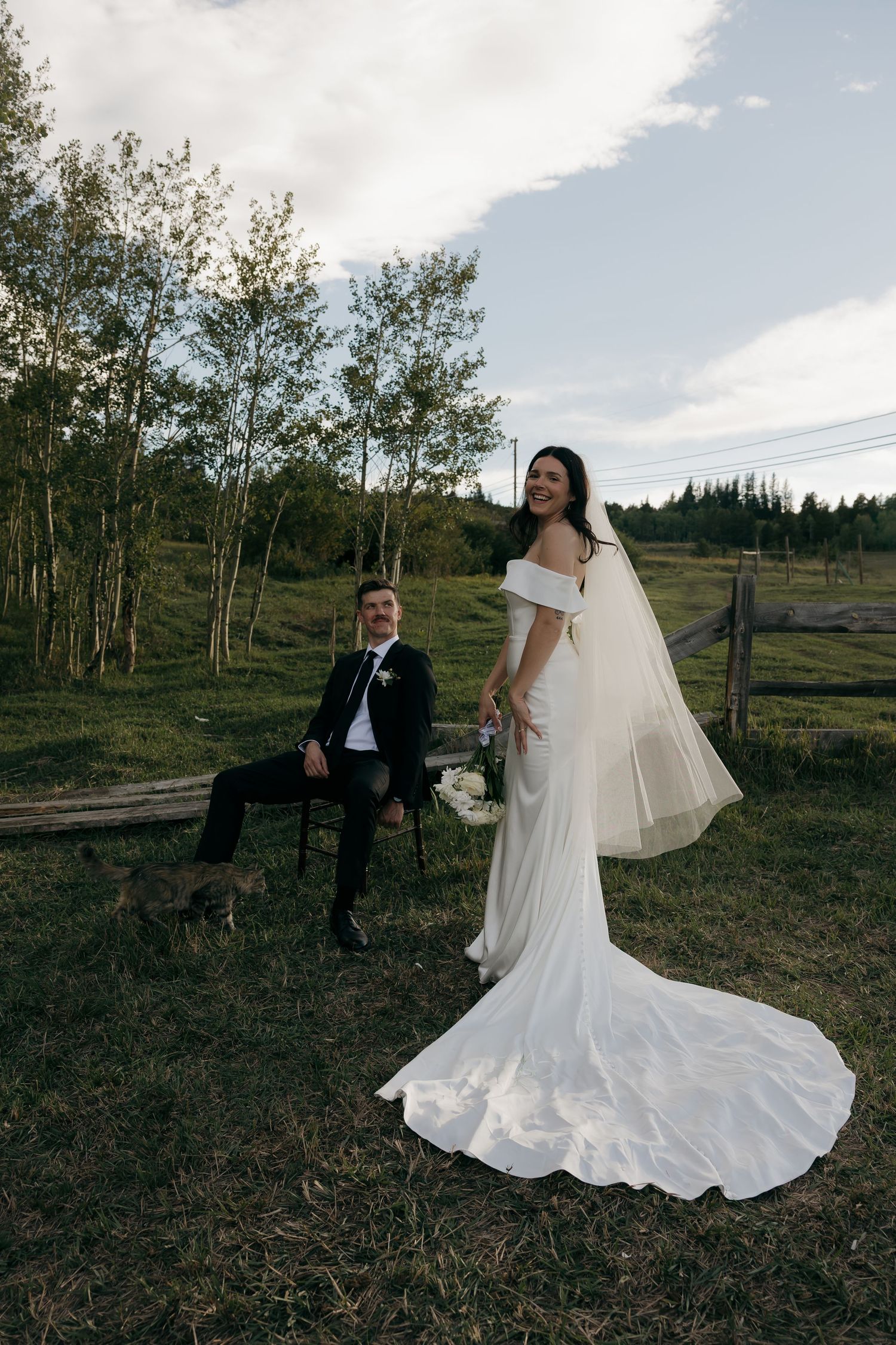 Bride in off-shoulder gown with veil poses while groom sits on chair outdoors.