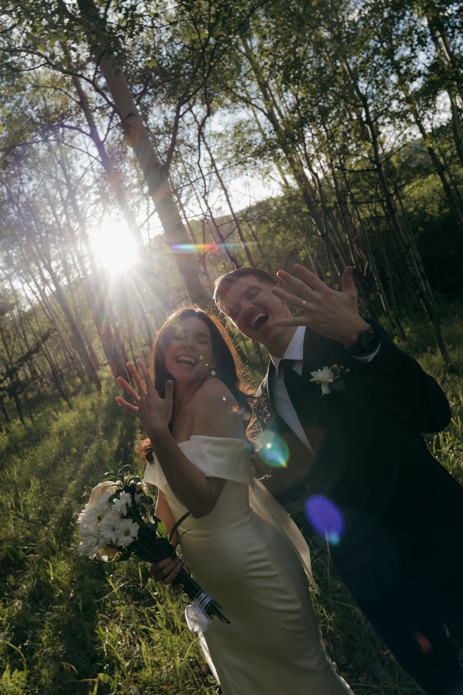 Joyful newlyweds showing wedding rings in sunlit birch forest after ceremony.