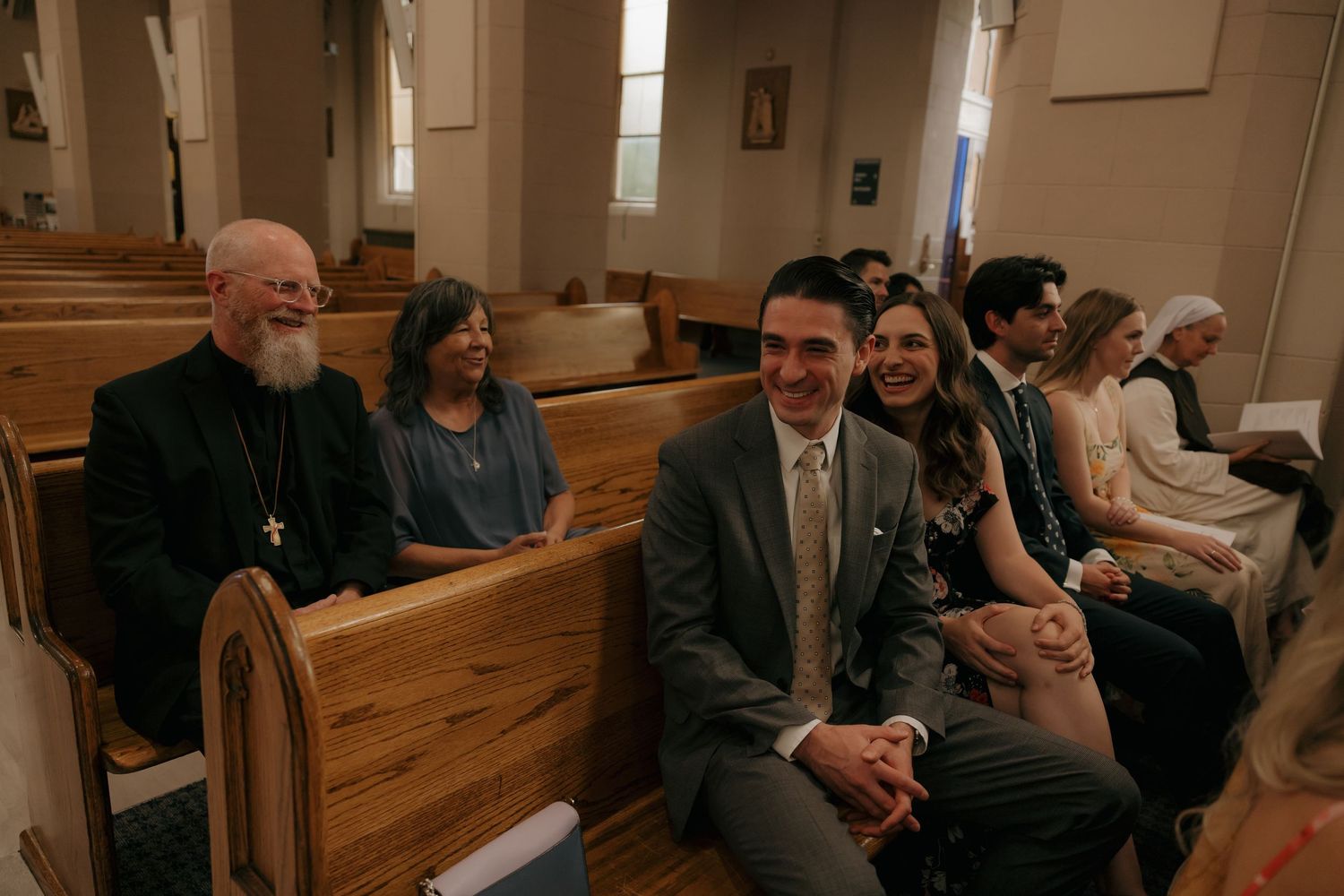 Smiling wedding guests including clergy seated in church pews during Catholic wedding ceremony.