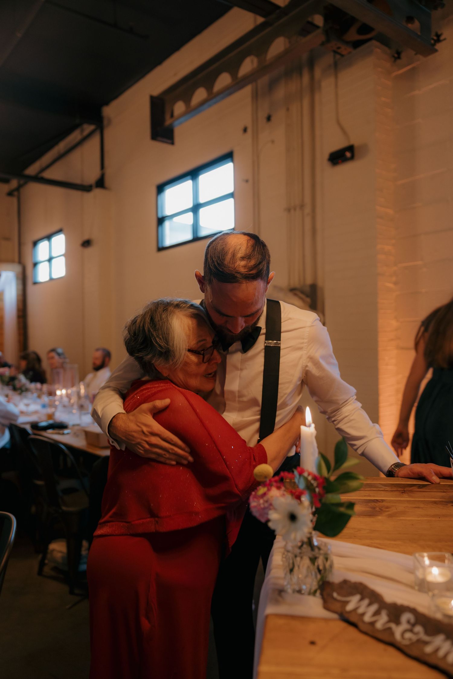 Groom in white shirt and suspenders embracing elderly woman in red dress at wedding reception.