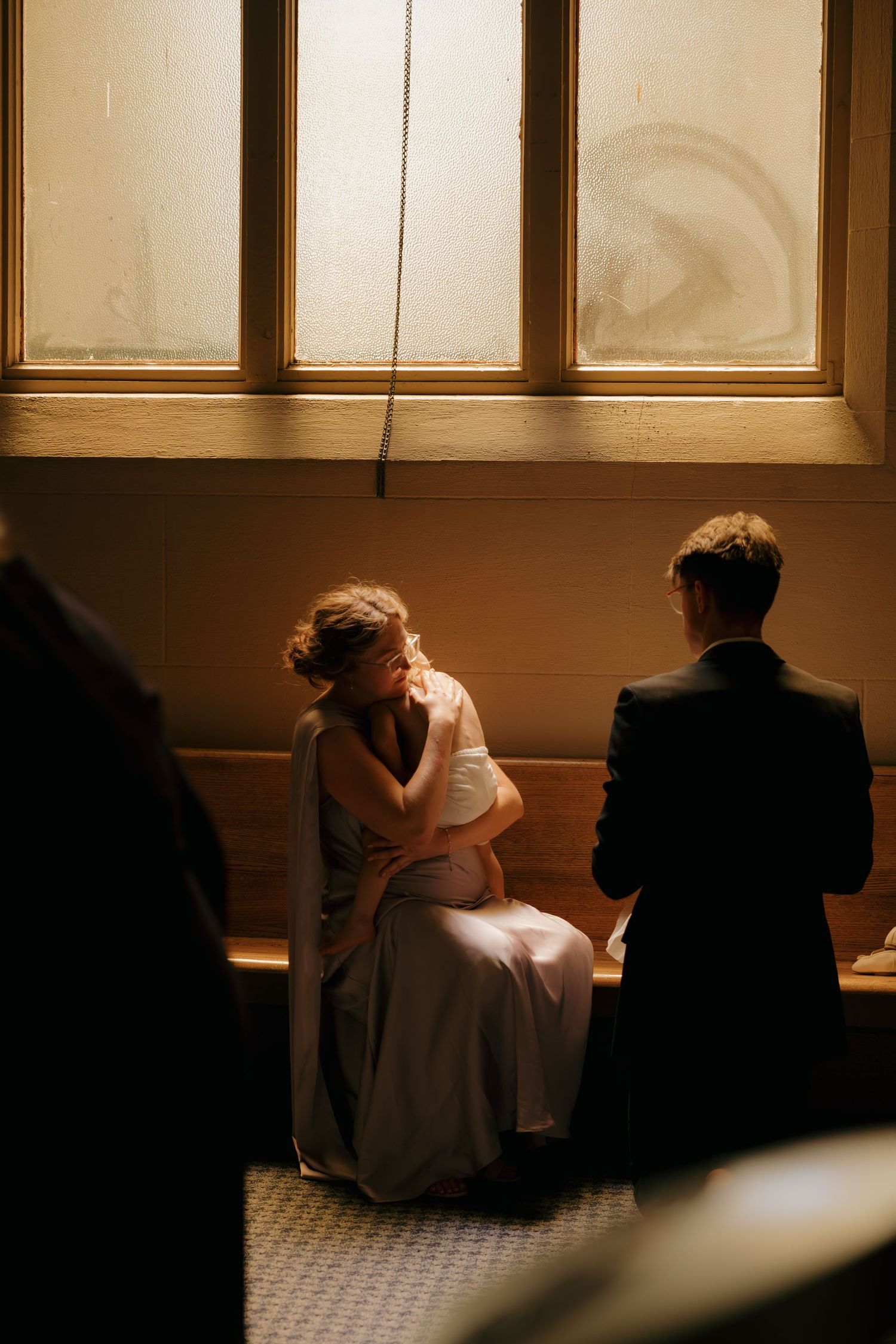 Bride in white gown sits pensively on bench beside groom in dark suit, bathed in warm window light.