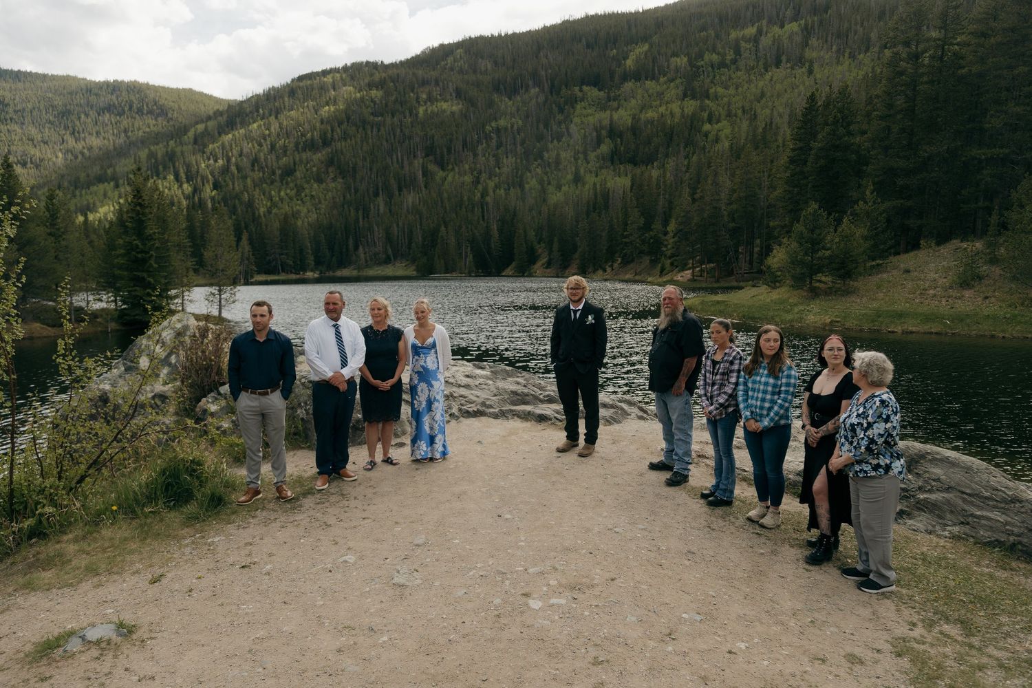 Group of people posing near a mountain lake surrounded by pine forests on a rocky path.