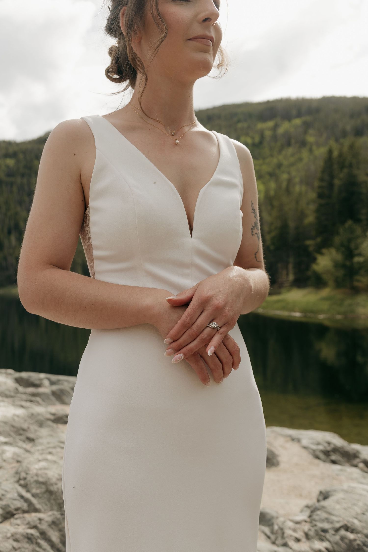 Bride in elegant white deep V-neck wedding gown clasping hands near a mountain lake with lush forest backdrop.