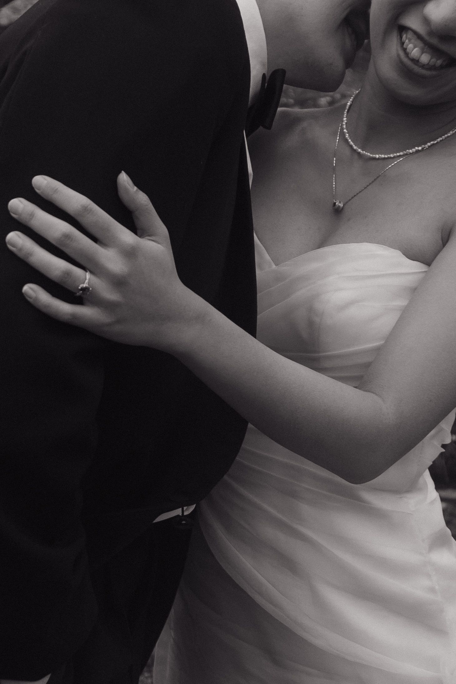 Black and white close-up of bride showing engagement ring while embracing groom in a tuxedo.