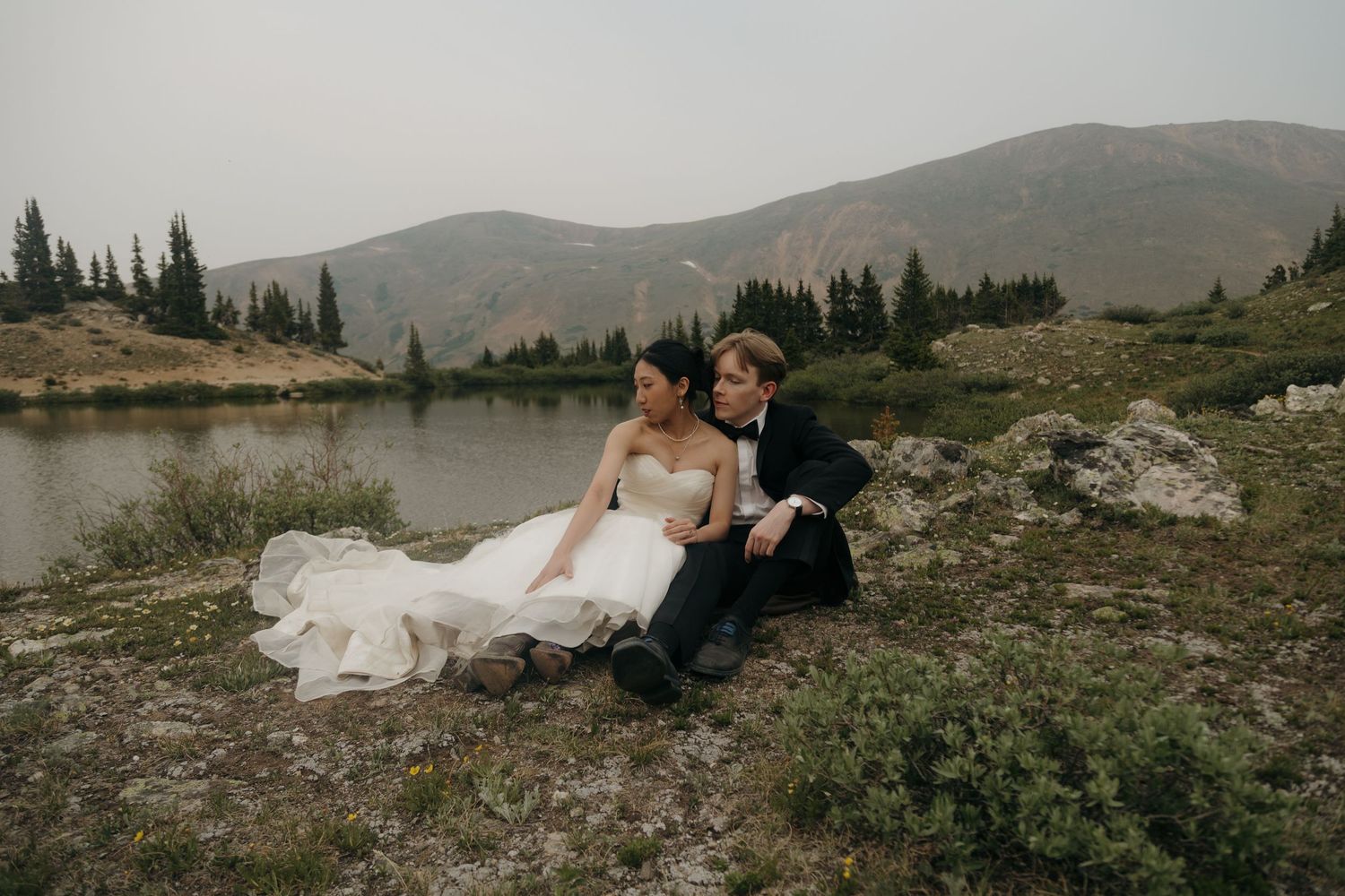 Bride and groom sitting together on rocky mountain shore in wedding attire during elopement session.